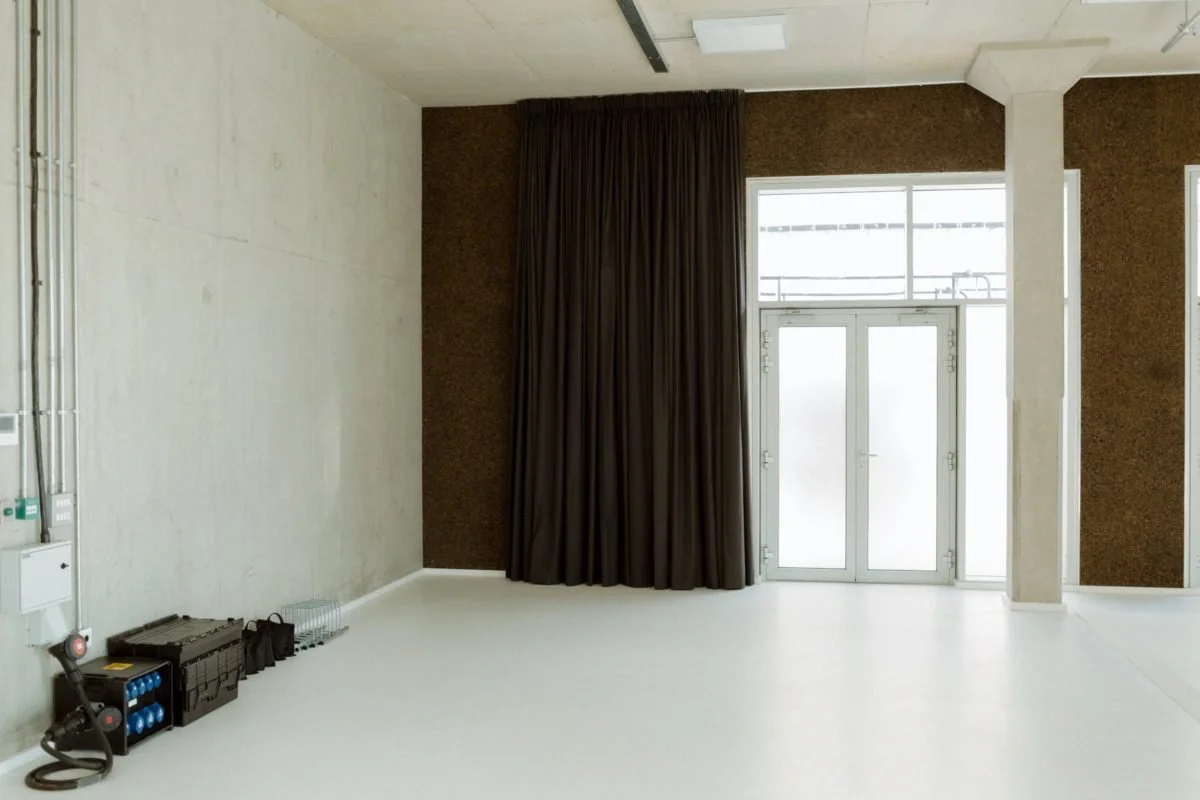 Empty room with white walls, a dark curtain covering a window, and a glass door leading outside. There are some equipment and bags on the floor near the wall.