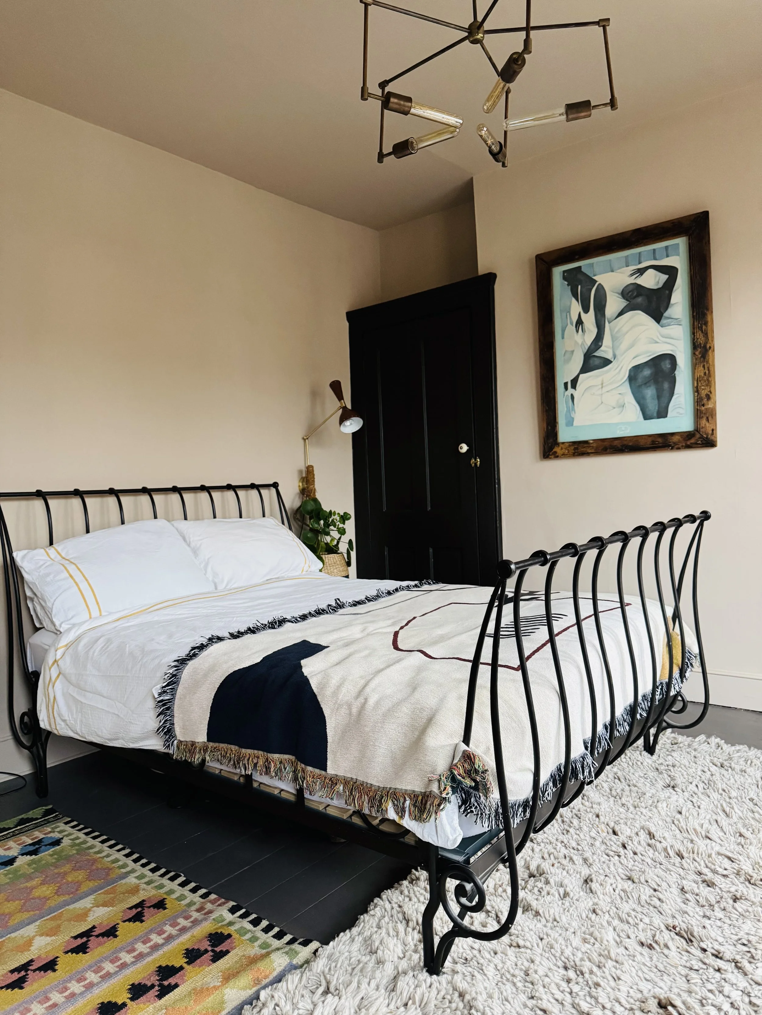 A bedroom with a black metal bed frame, white bedding with yellow accents, a fringed blanket, a colorful rug, a white shaggy rug, a black wardrobe, a wall-mounted lamp, a modern pendant light fixture, a framed art piece of two women, and a small plan