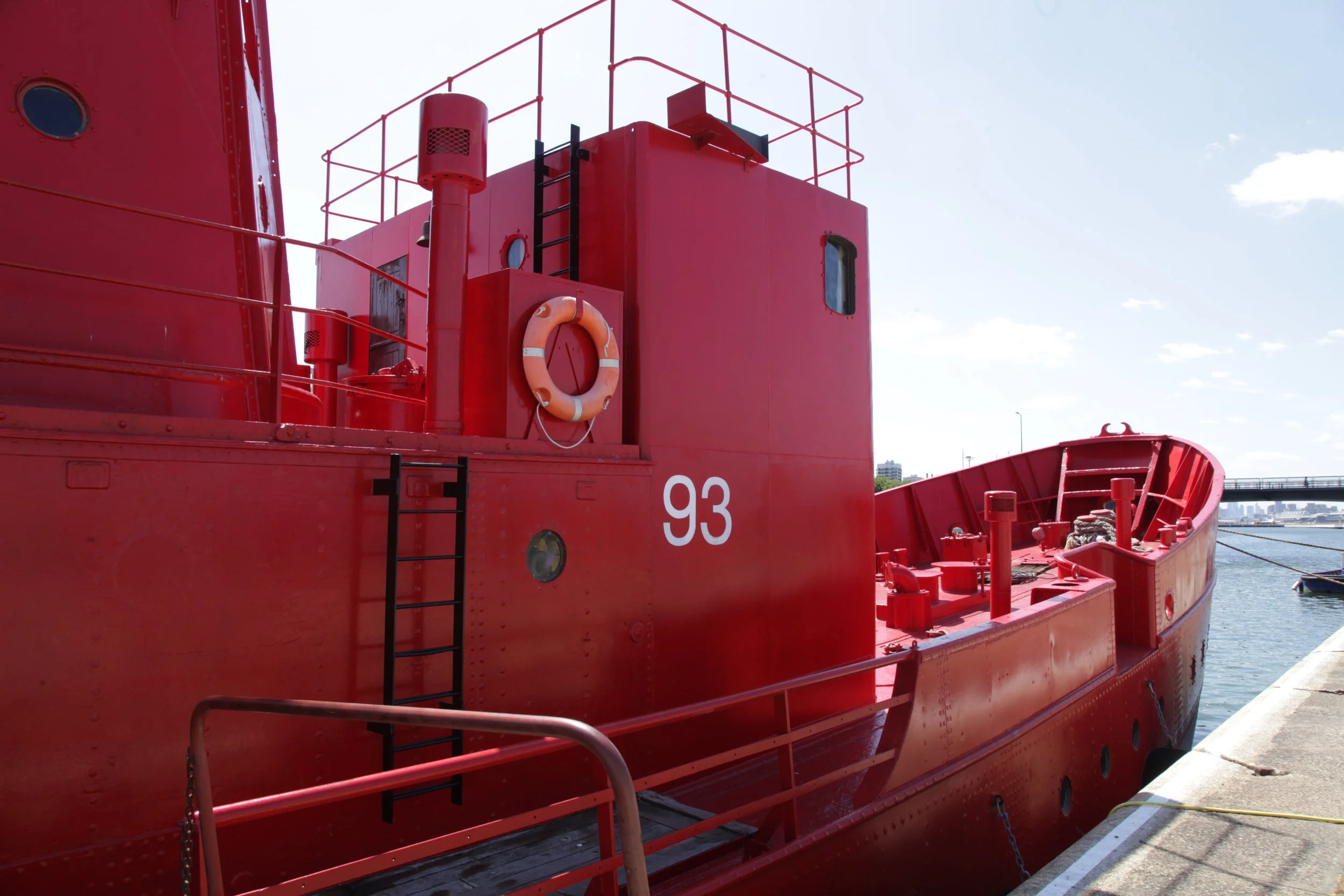 Close-up of a red submarine docked at a pier, with the number 93 on its side, featuring ladders, a life ring, and safety equipment.
