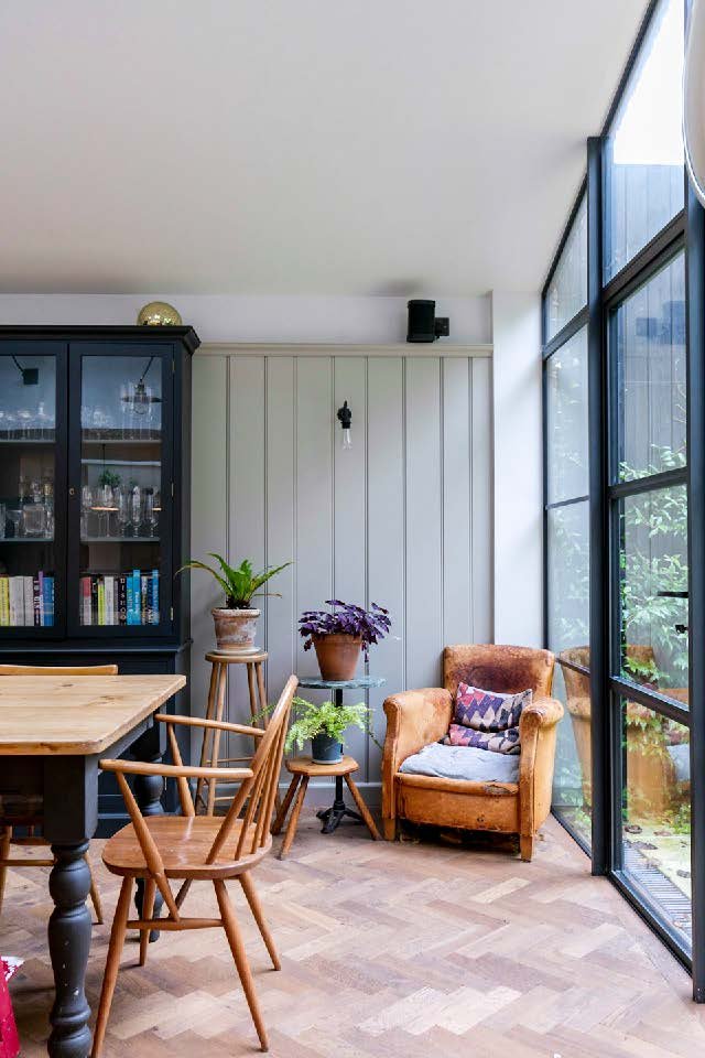 A cozy dining corner with a wooden table and chairs, a black cabinet with glass doors, green plants on small tables, an orange armchair with a cushion, and large glass sliding doors leading outside.