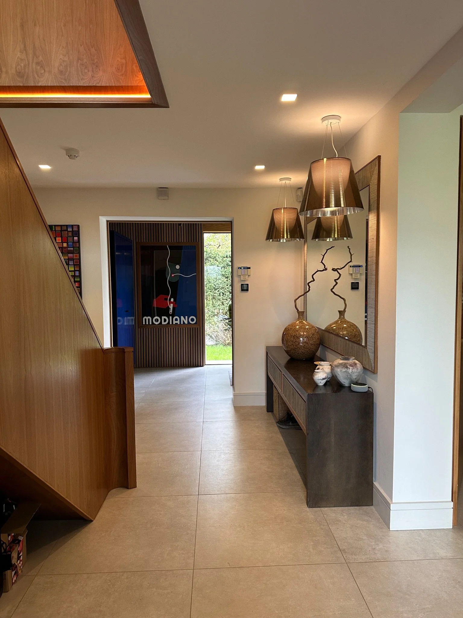 Modern entrance lobby with wooden staircase, decorative vases, large mirror, pendant lights, and a door with colorful artwork and glass panel.