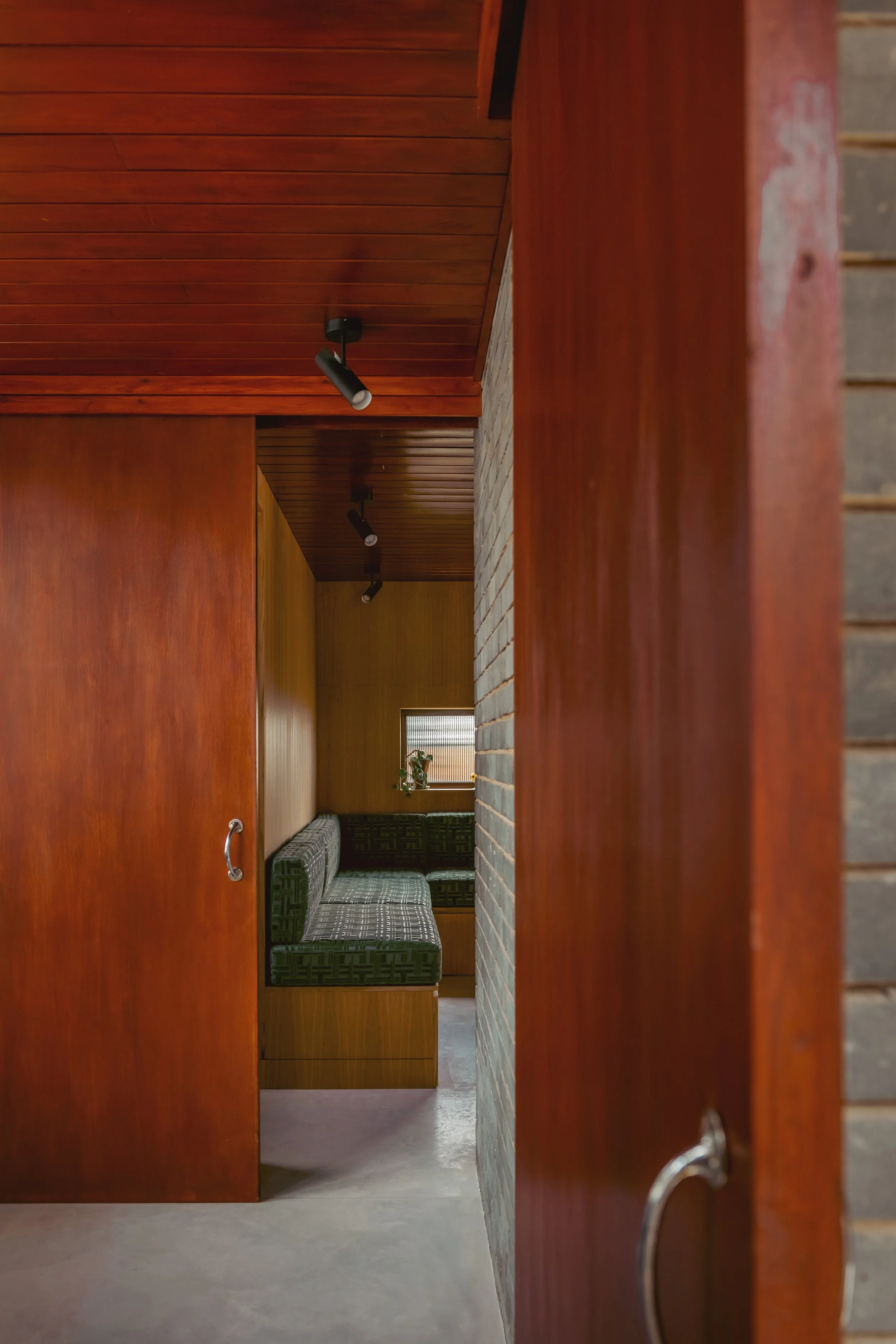 View through a partially open wooden door into a cozy corner with a green patterned bench, small window with a potted plant, and wood-paneled walls and ceiling, illuminated by spotlights.