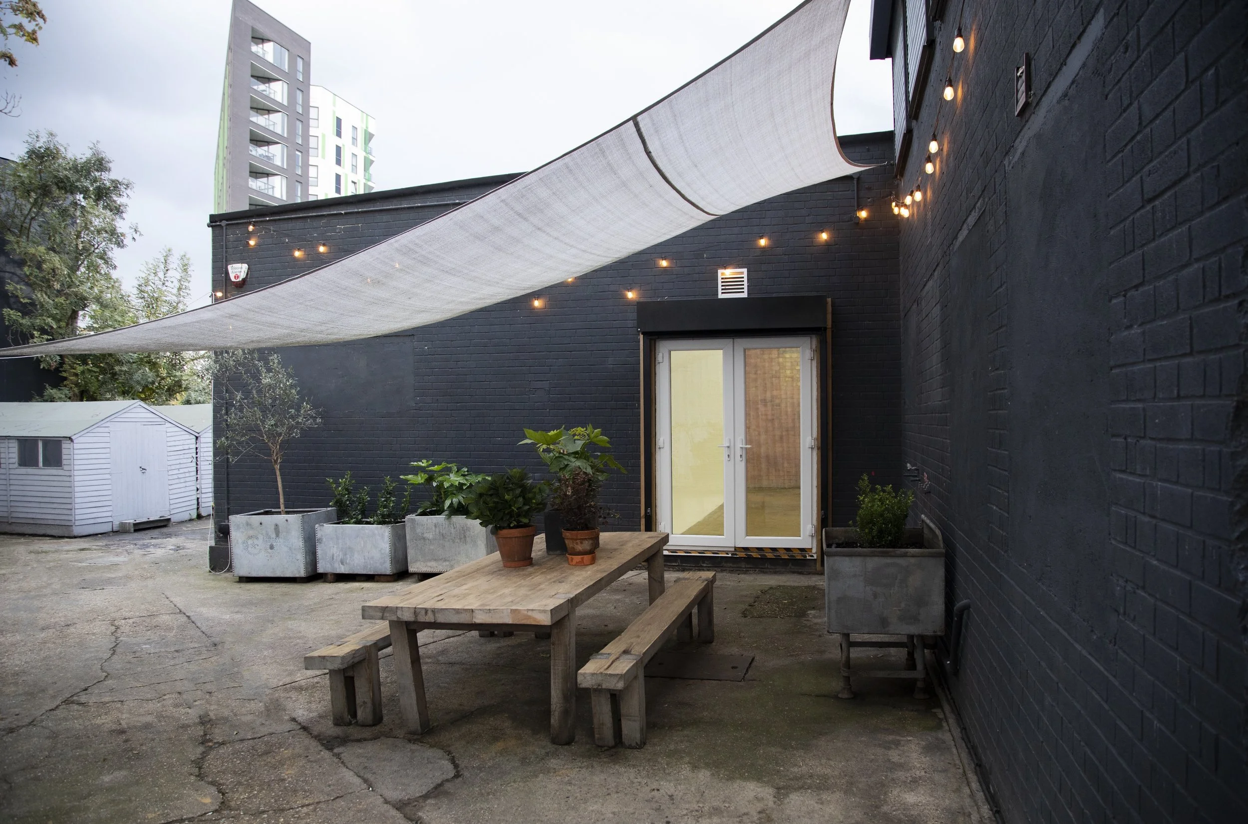 An outdoor backyard area with a black brick building, string lights, potted plants, a wooden picnic table with benches, a white shade sail overhead, and a small white shed in the background.