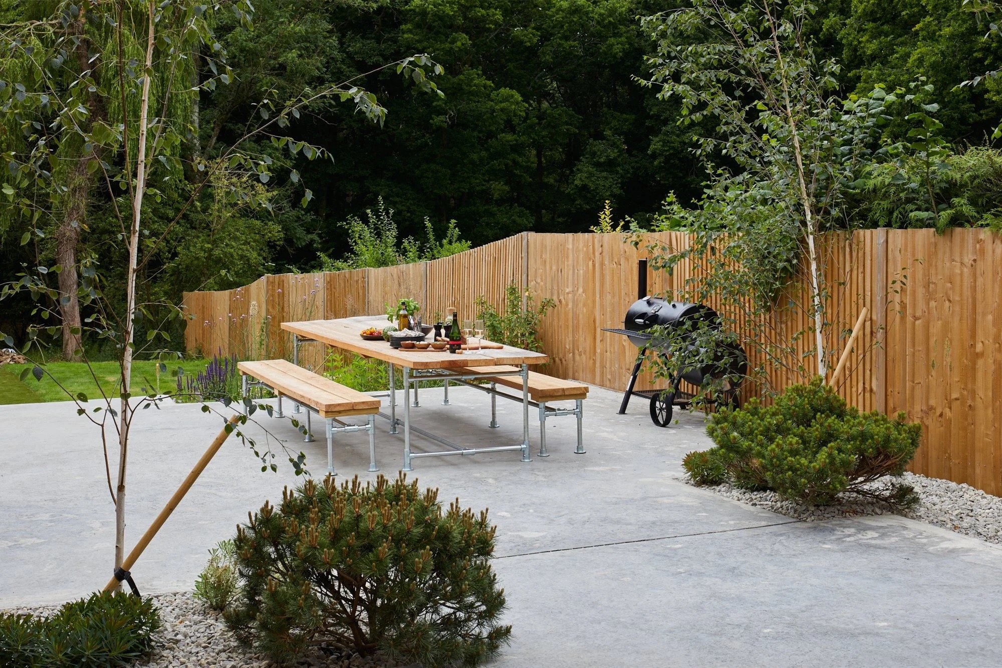 A backyard patio with a wooden picnic table and benches set for a meal. There is a charcoal grill near a wooden fence, surrounded by green trees and shrubs.