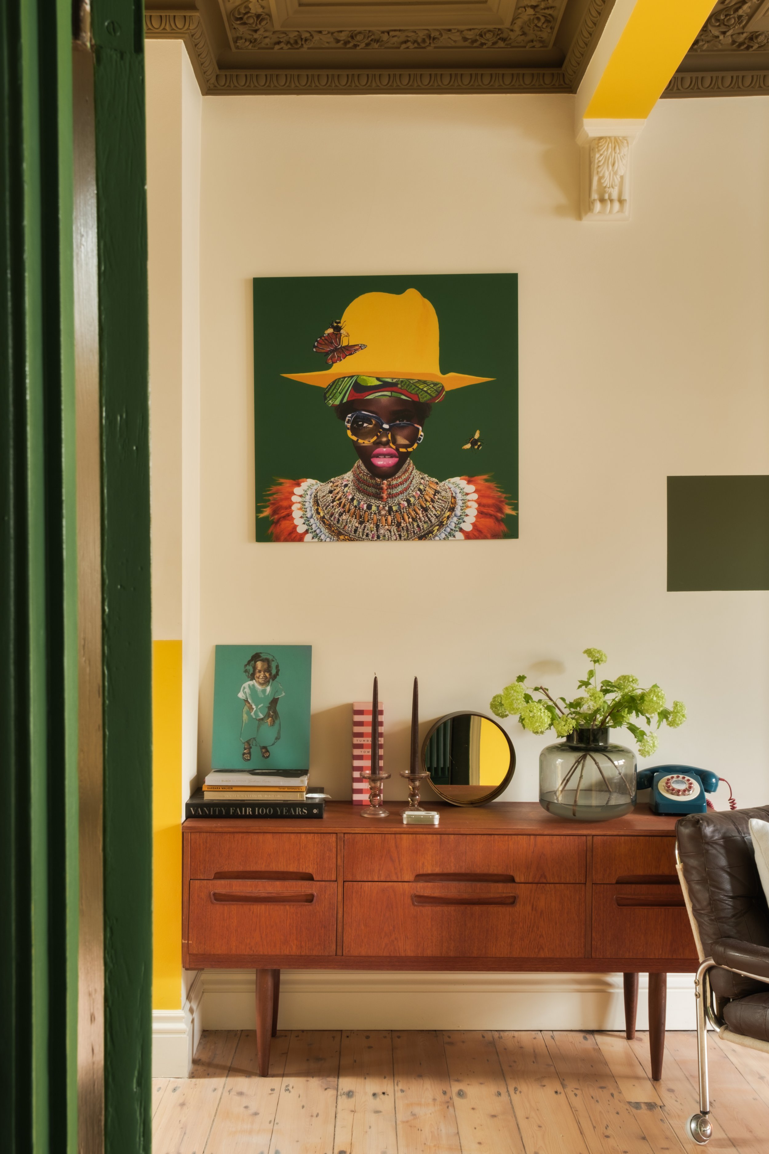Interior of a room with a wooden sideboard, decorative items, a large colorful artwork of a woman with a large yellow hat on the wall, and a vase with green flowers.