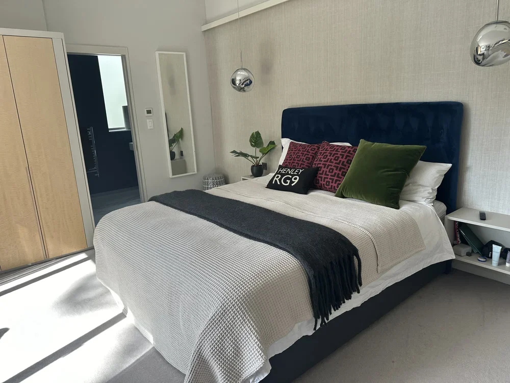 Modern bedroom with a large bed, navy blue upholstered headboard, white bedding, and decorative pillows, including a green velvet pillow and a black pillow with white text. There are two silver hanging pendant lights above the bed, a small white nigh