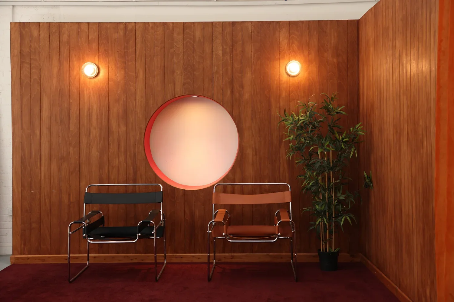 Interior of a waiting area with two chairs, a potted plant, and warm lighting on a wood-paneled wall.