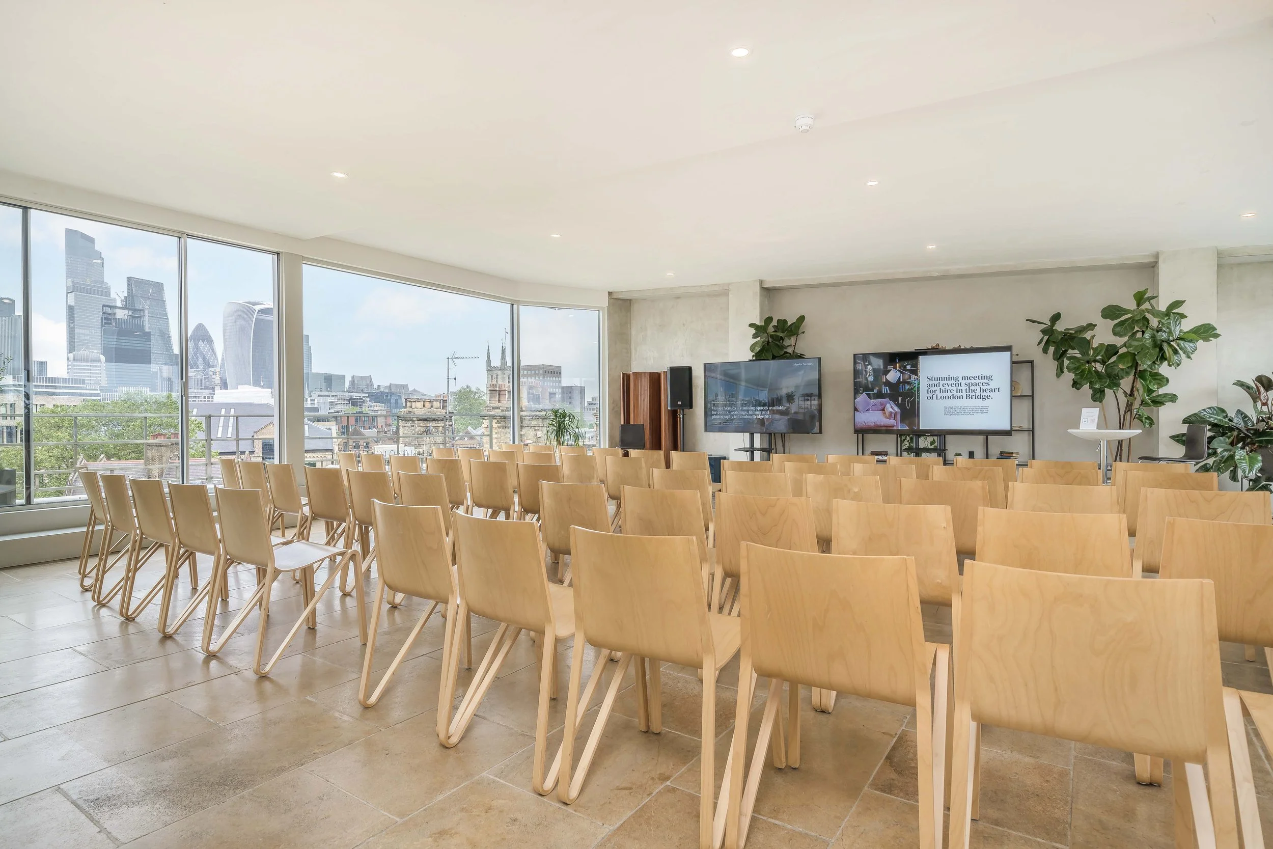 Conference room with rows of wooden chairs, large windows with city skyline view, and two screens at the front for presentations.