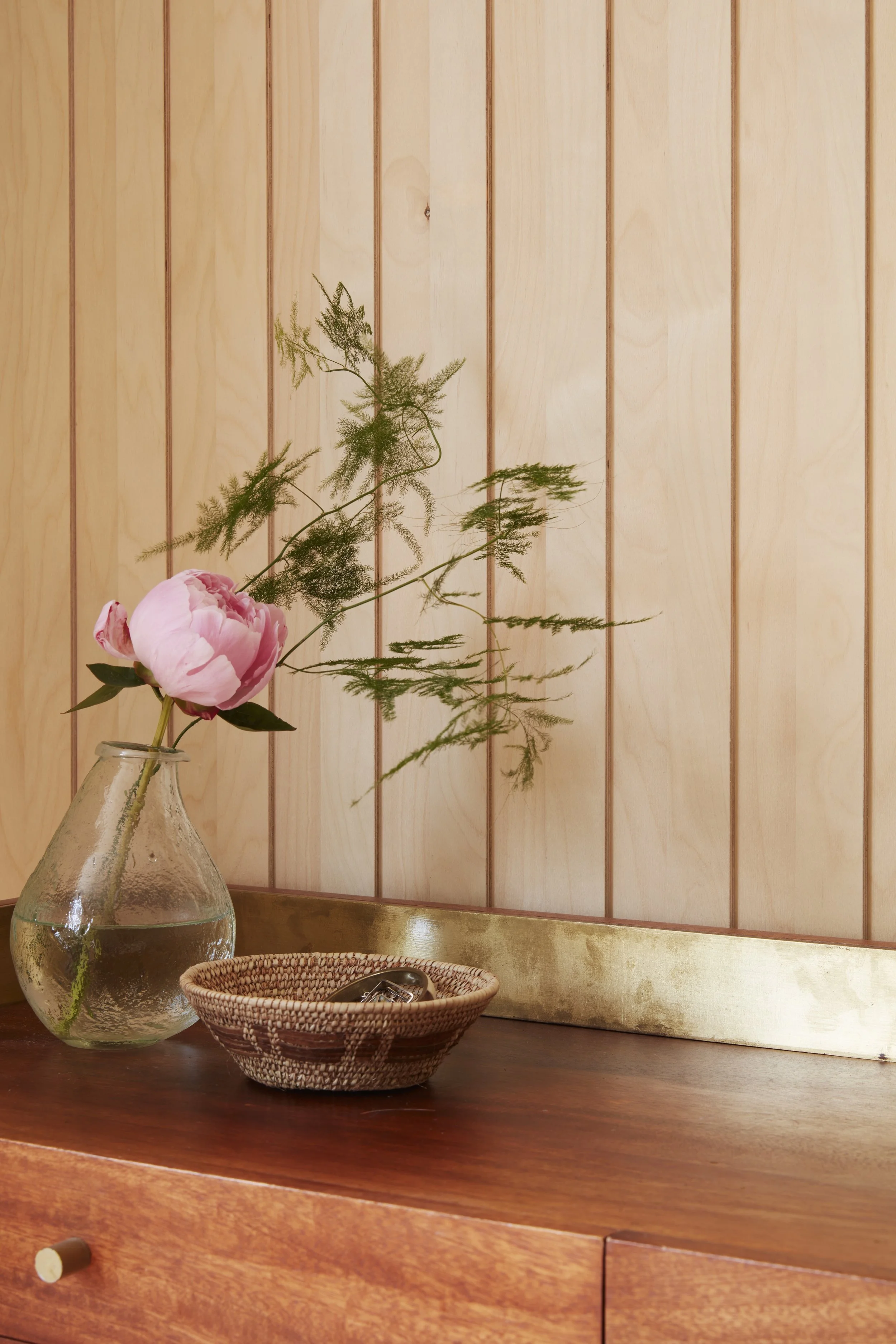 A vase with a pink peony and green greenery, a woven basket with a pair of eyeglasses, on a wooden surface against a wood-paneled wall with a brass trim.