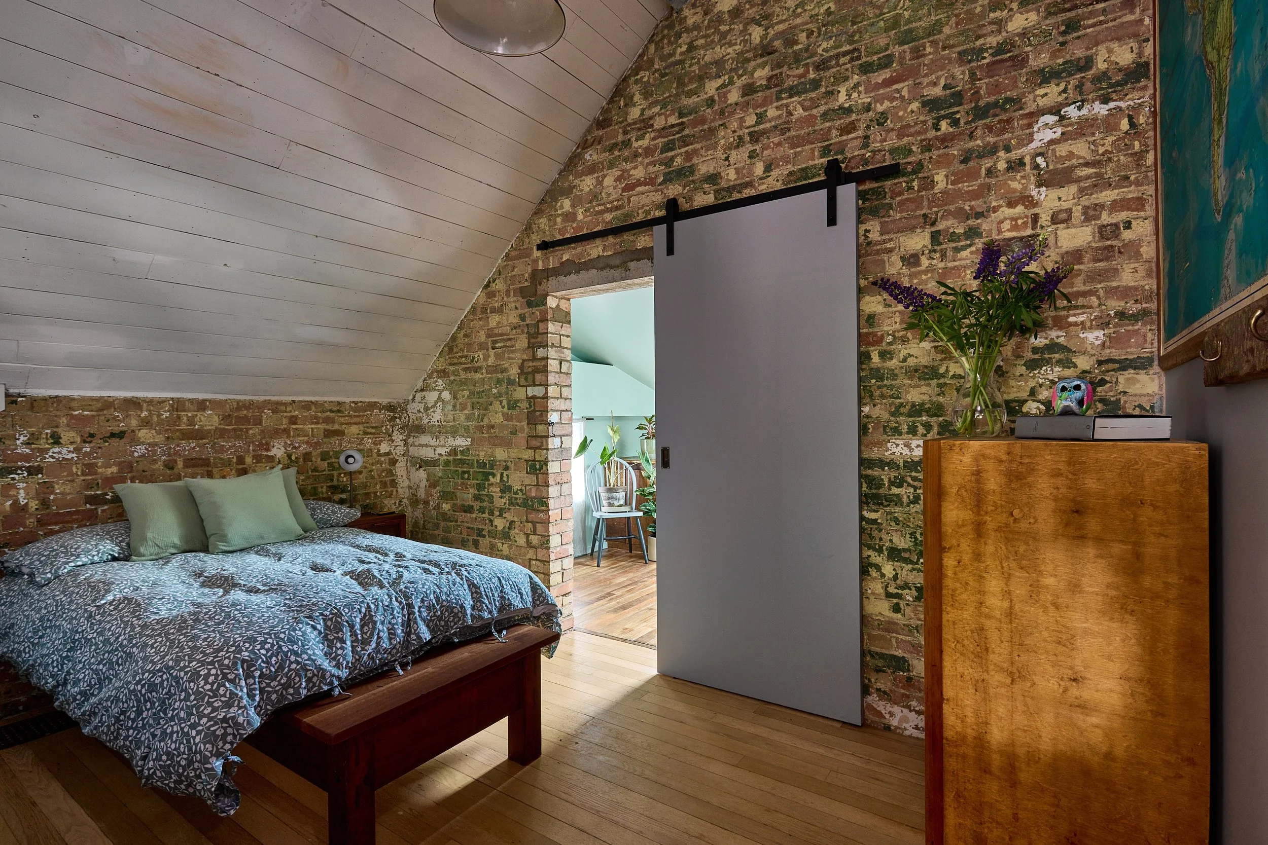 Bedroom with exposed brick wall, wooden floor, bed with gray and white patterned bedding and green pillows, sliding gray door, wooden dresser with purple flowers, small decorative owl, and a picture hanging on the wall.