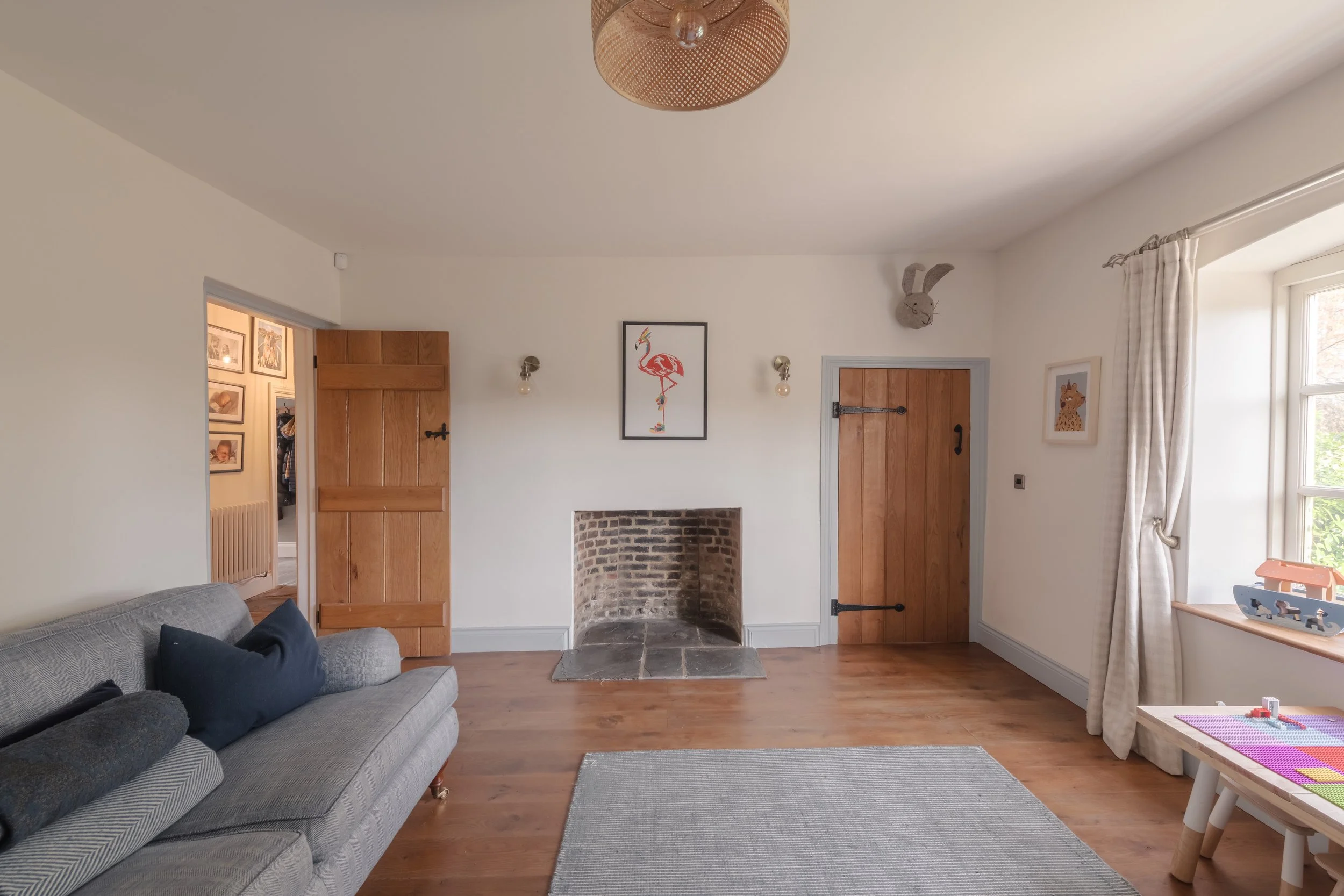 Living room with a grey sofa, wooden floors, a fireplace, and decorated with children's artwork and stuffed animals, with a window on the right.