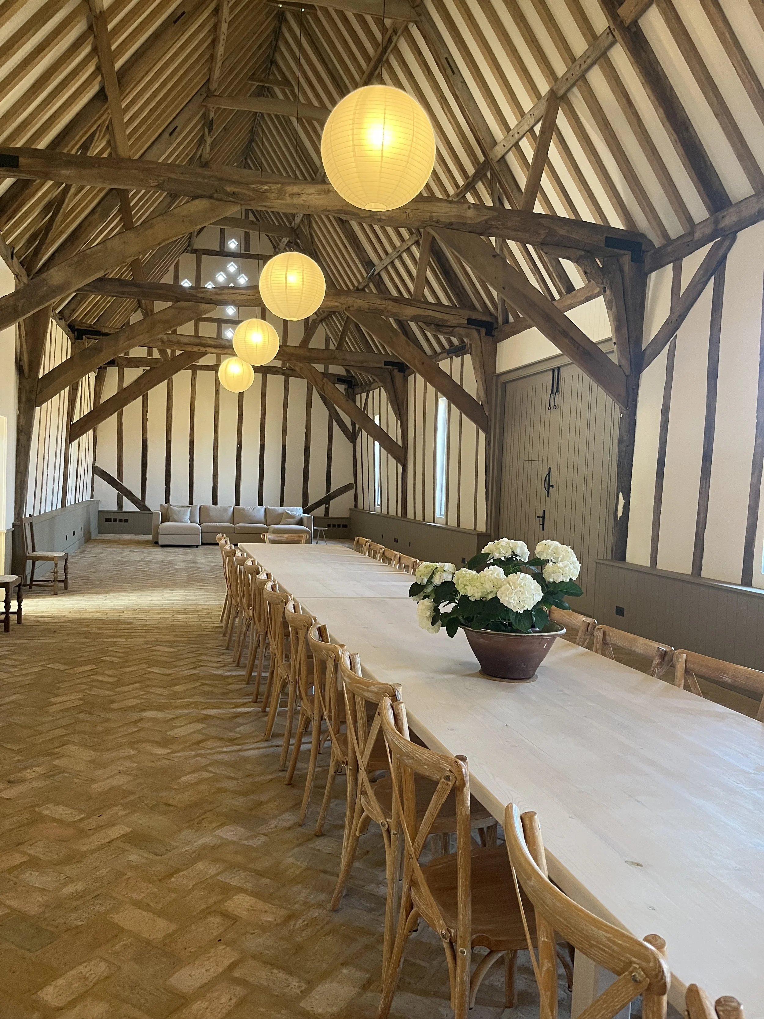 Long wooden table with chairs on either side, large pot with white hydrangea flowers in the center, rustic barn interior with wooden beams and paper lantern ceiling lights.