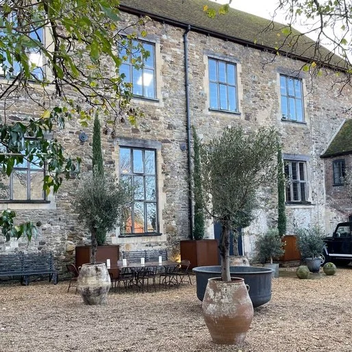 Stone building with several tall windows, plants in pots, outdoor furniture, and a black vehicle parked nearby.