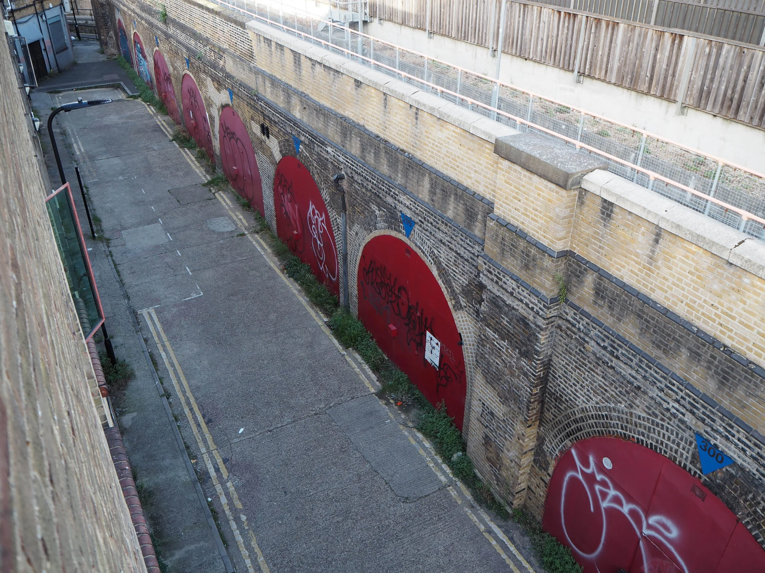 An alleyway with a concrete pavement, graffiti-covered red doors in a brick wall, street signs and a lamppost.