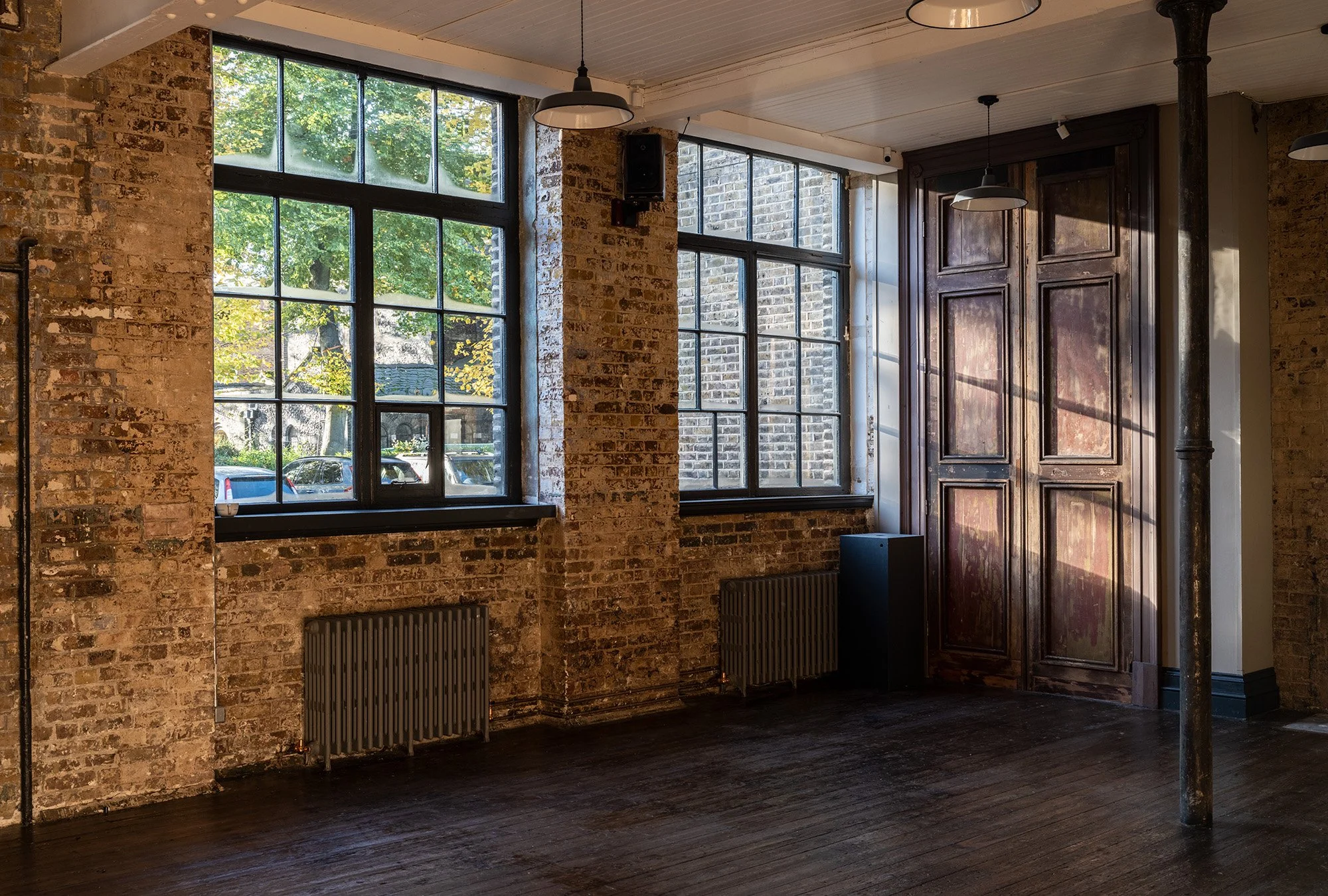 Interior of a room with large industrial-style windows, brick walls, a wooden door, and dark wooden floors, illuminated by natural light.