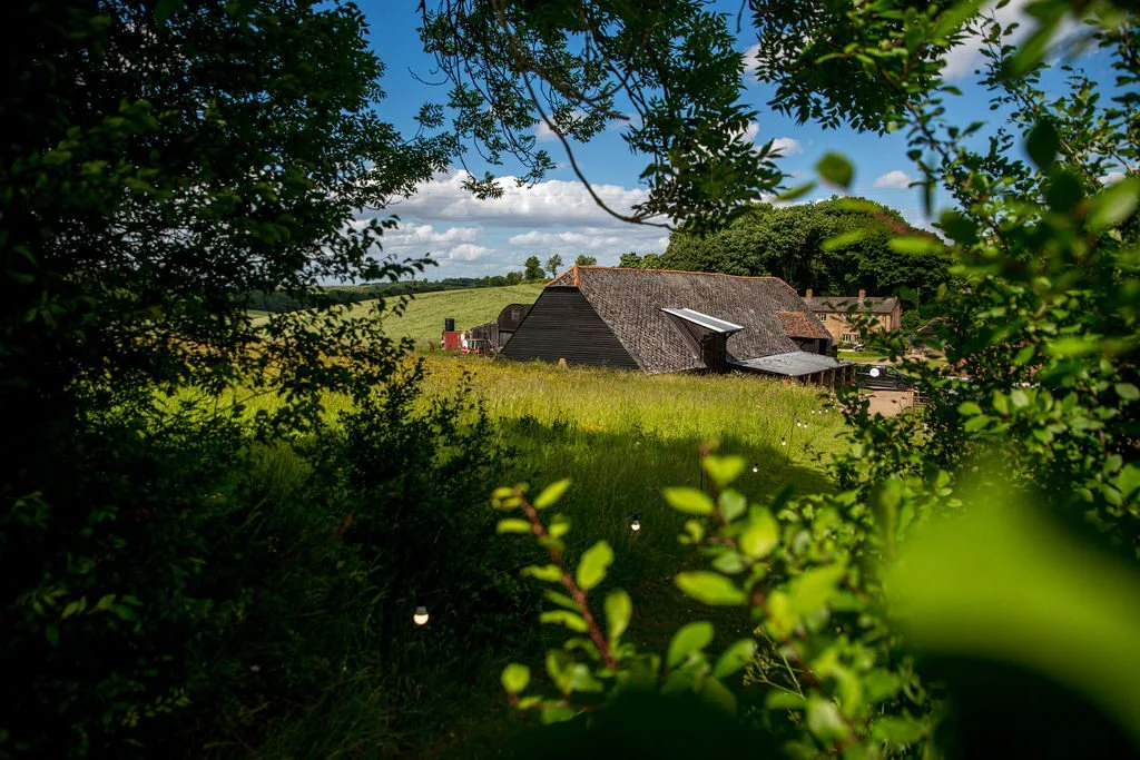 A rural farm scene with a barn and other buildings surrounded by green grass and trees, under a partly cloudy sky.