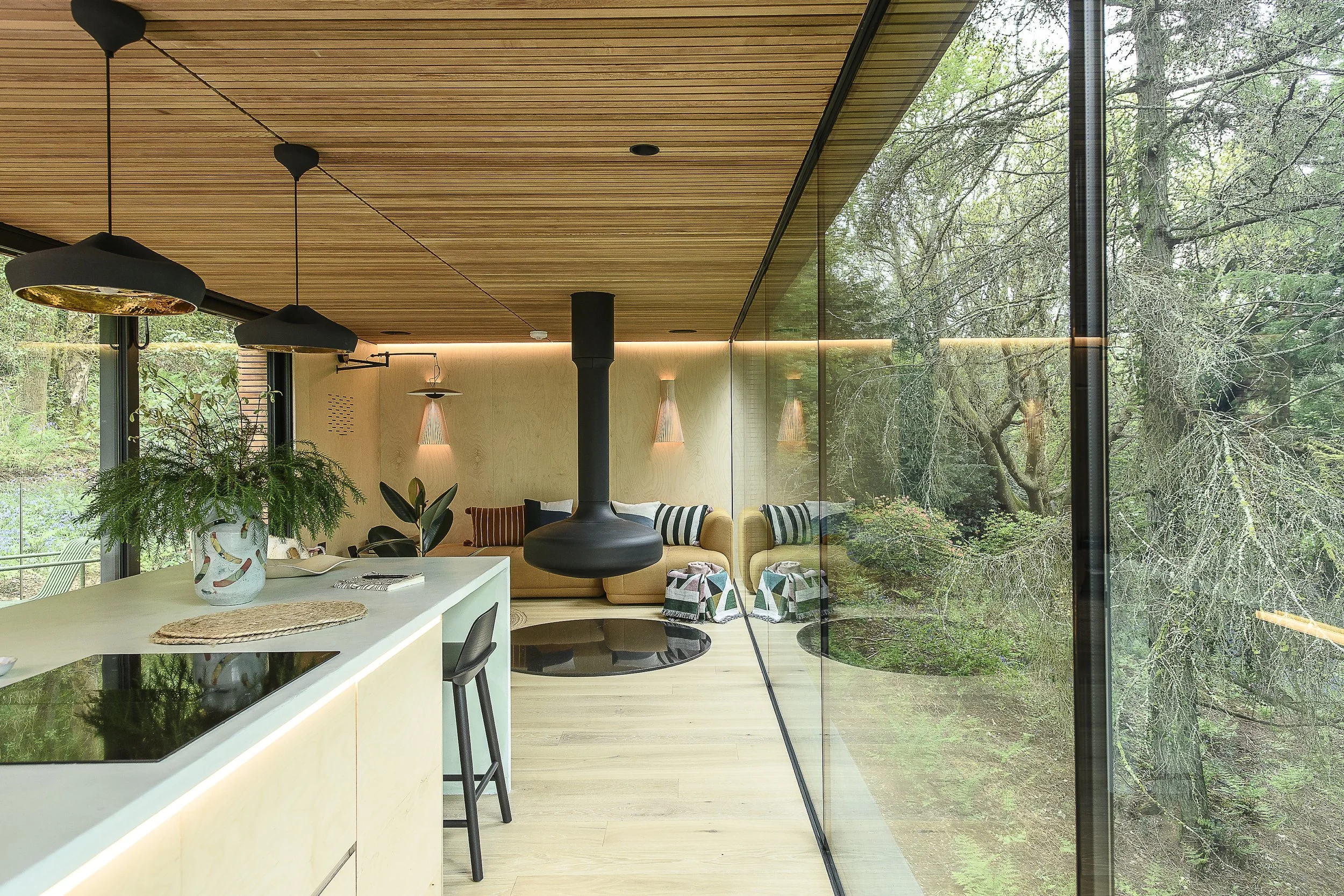Modern living room with wood-paneled ceiling, large glass wall overlooking a lush forest, a beige sectional sofa with striped cushions, hanging pendant lights, a black fireplace, and an open kitchen island with a potted plant.