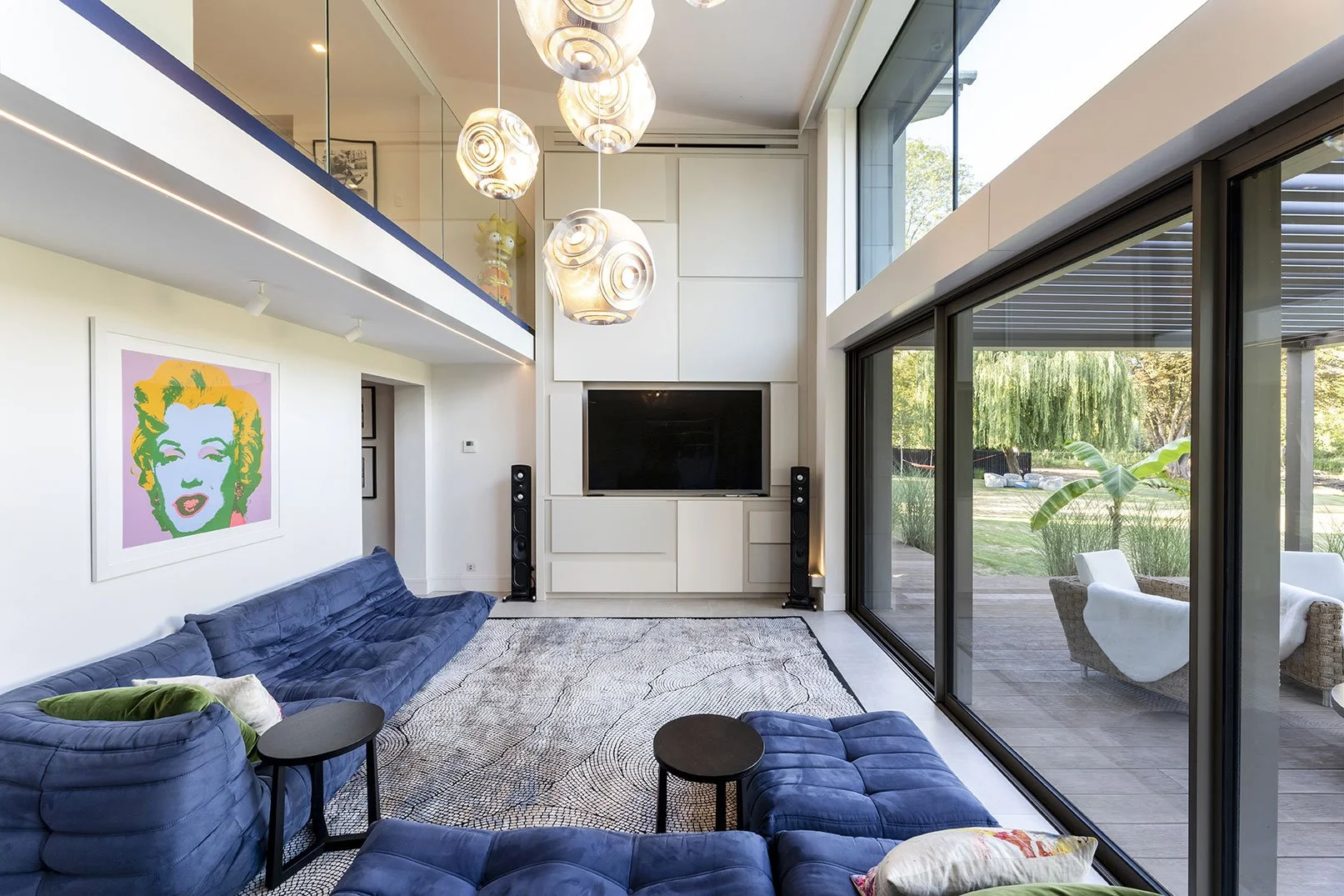 Modern living room with a blue velvet sectional sofa, abstract framed artwork of Marilyn Monroe on white wall, large flat-screen TV, black tower speakers, glass sliding doors opening to outdoor patio, and contemporary hanging light fixtures.