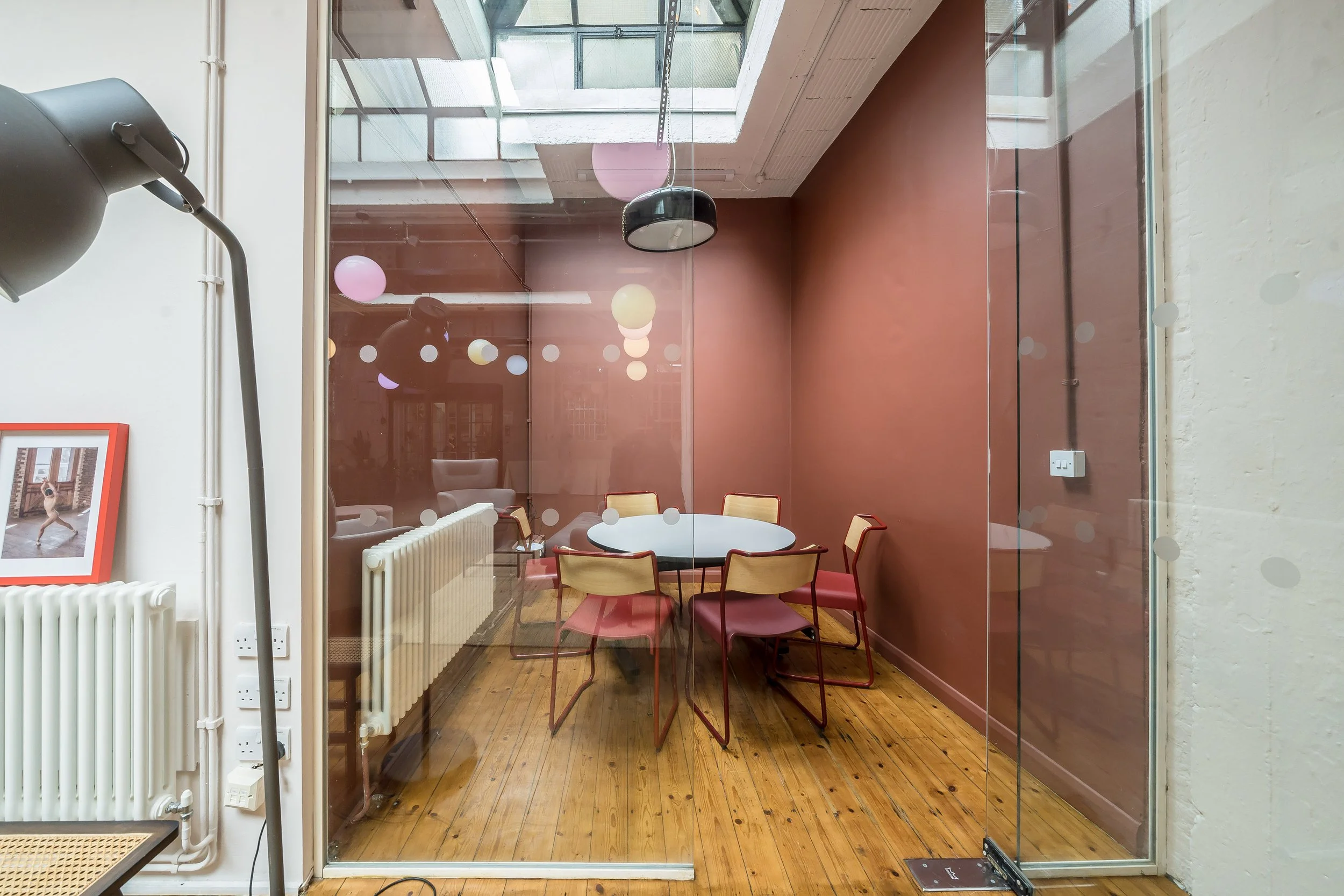 A small conference room with a round white table and six chairs, three with yellow backs and pink seats, enclosed by glass walls. The room has a red wall, wooden floor, and a black hanging lamp. Outside the room, there is a white wall with a radiator