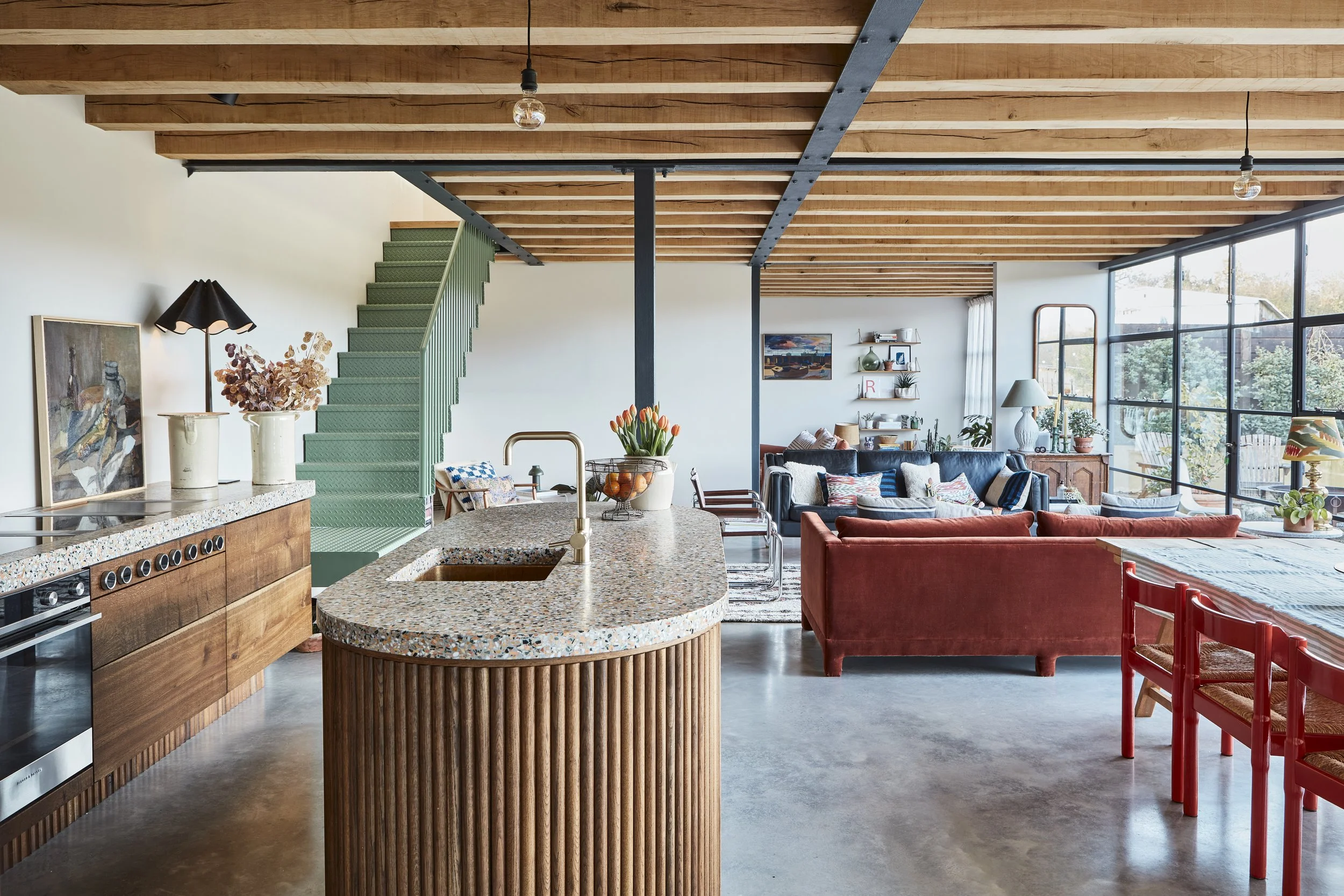 Open-concept living room and kitchen with wooden ceiling beams, large windows, a red sofa, a variety of plants, and decorative items.