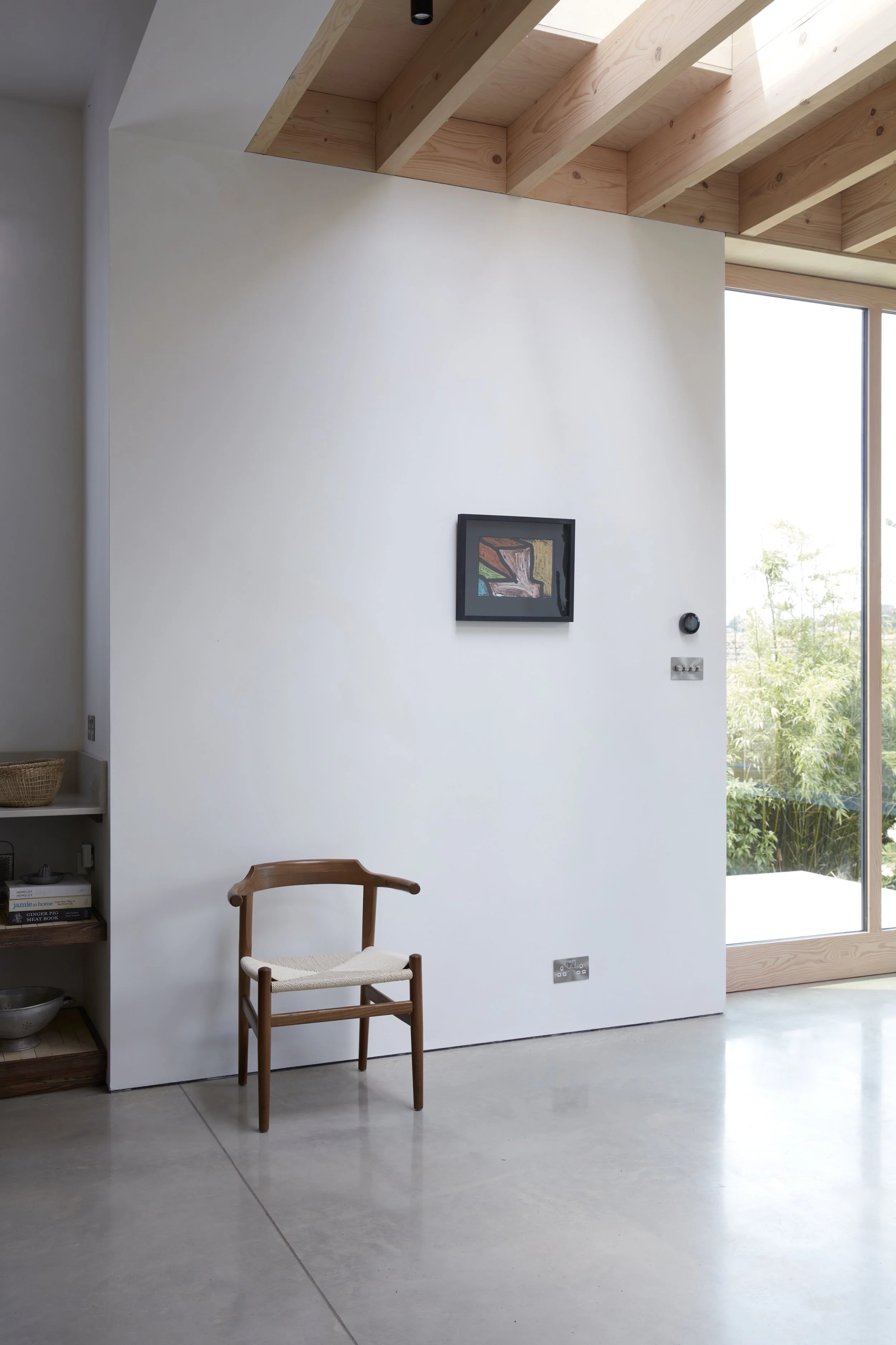 A minimalist interior with a white wall, a small wooden chair with a woven seat, shelves with books and decorative items, a framed abstract artwork, and large sliding glass doors showing greenery outside.