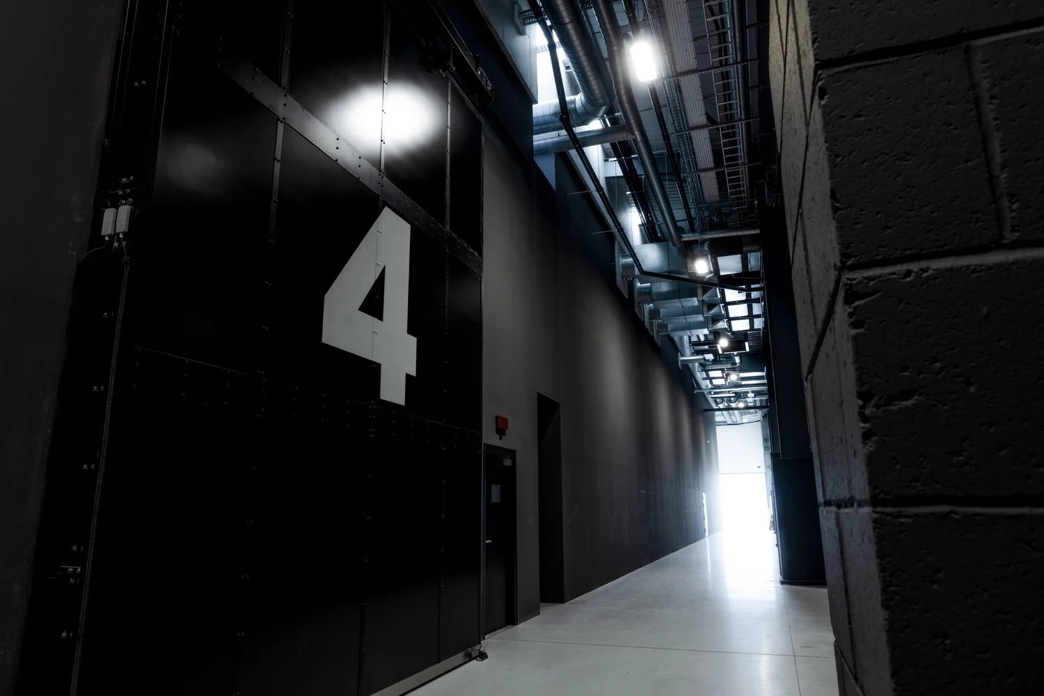 Industrial hallway with large black door labeled with the number 4, exposed ductwork and piping on ceiling, concrete block wall on the right, and light coming from an open area at the end of the hall.