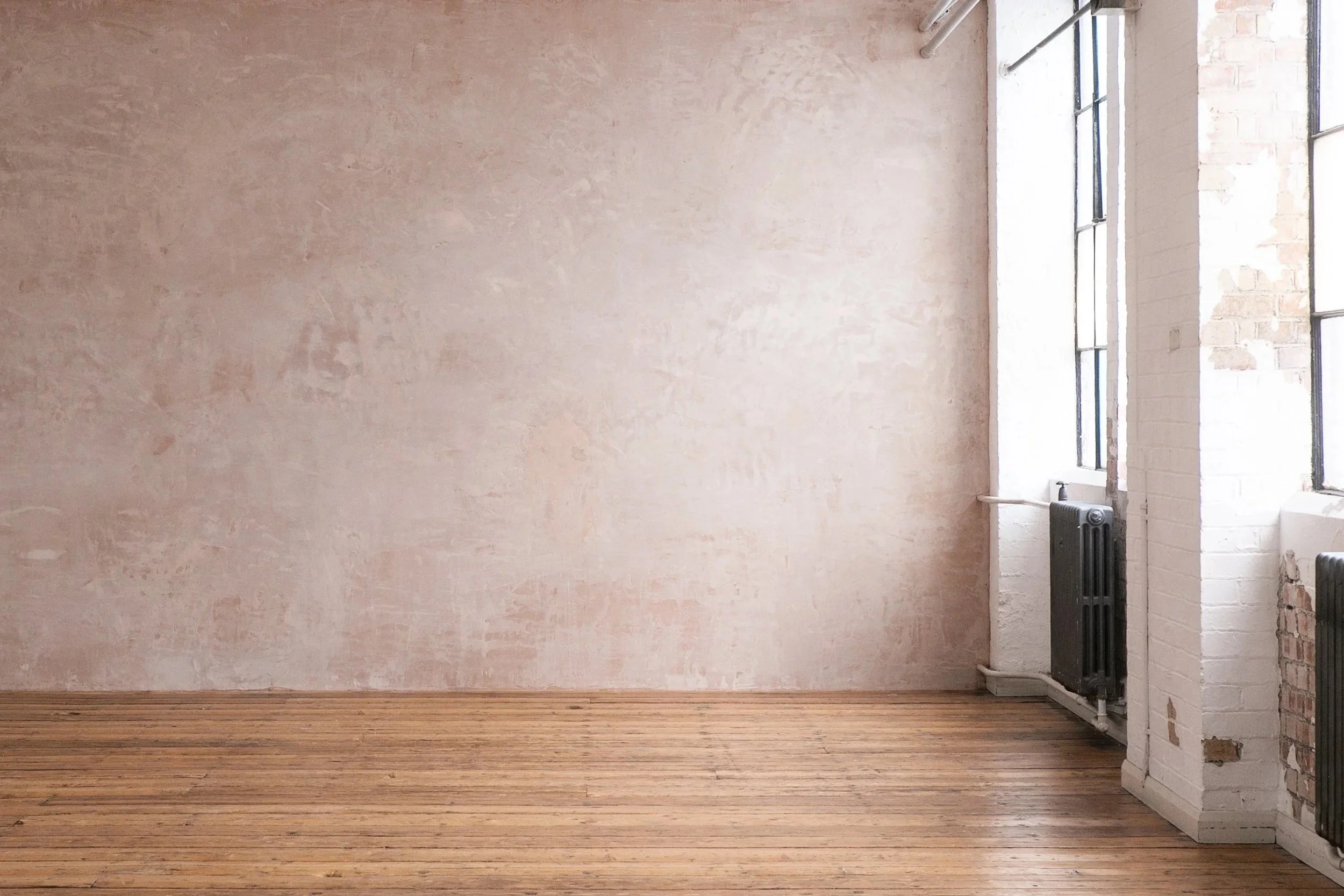 Empty room with light pink textured wall, wooden floor, large windows with black frames, and white painted brick wall with exposed bricks near the windows.