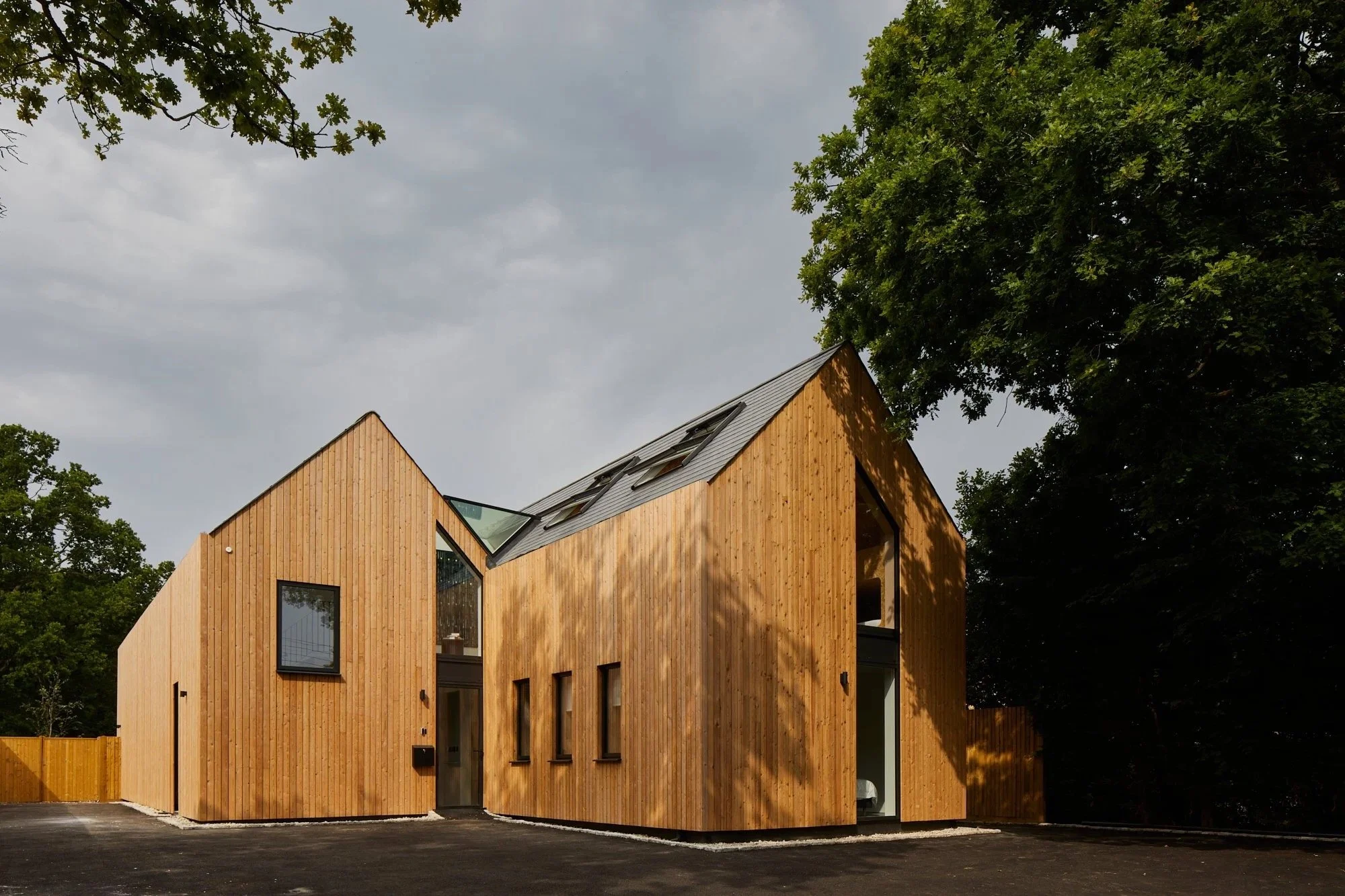 Modern house with wooden exterior walls, multiple windows, and a sloped roof, surrounded by trees and a paved driveway.