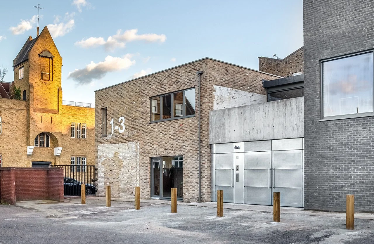 Modern brick building with large windows and metallic door, marked with the number 13, situated on a street with small parking spaces and wooden posts.