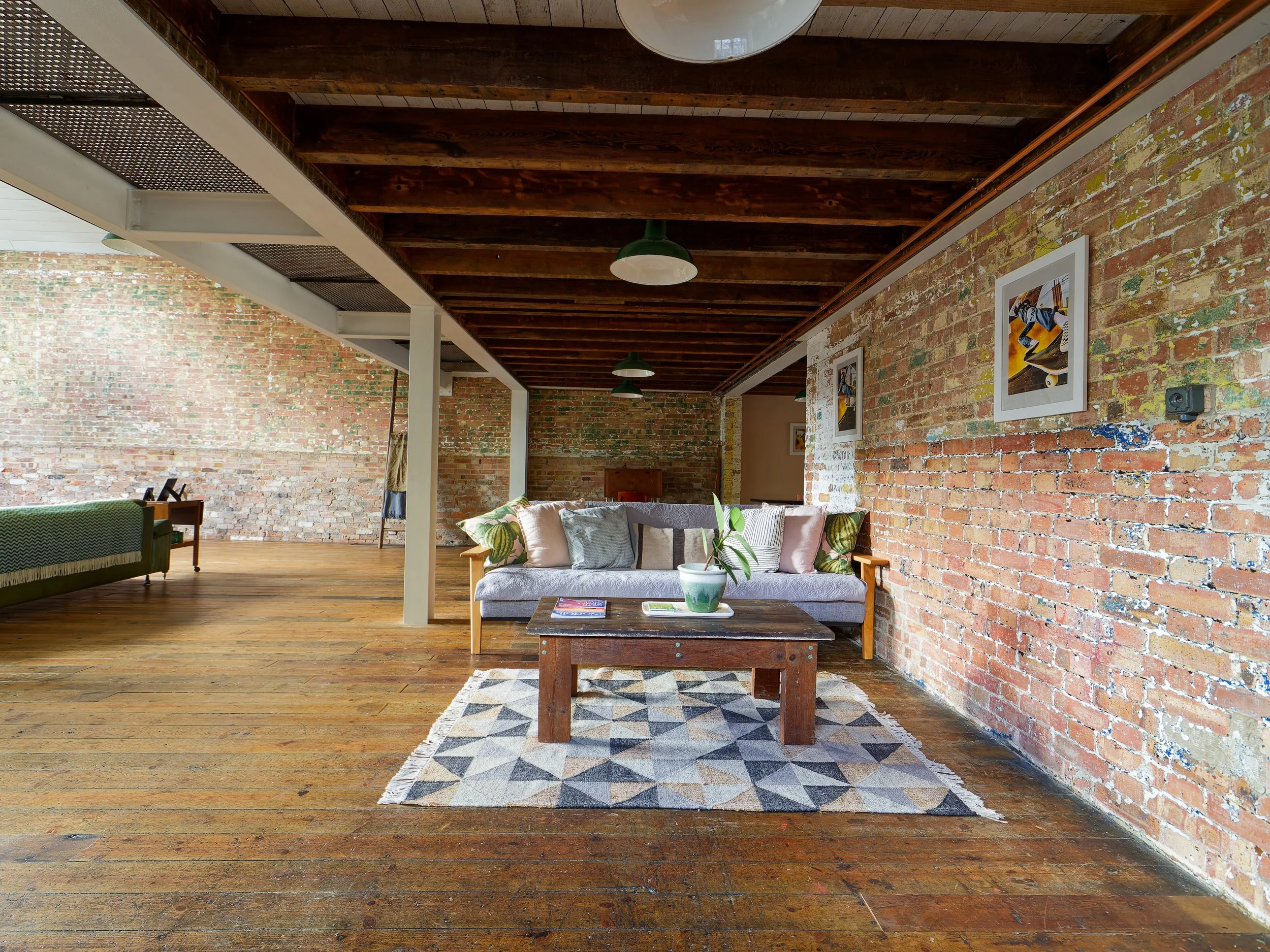A cozy living room with exposed brick walls, wooden floor and ceiling beams. There is a sofa with multiple pillows, a wooden coffee table with a plant and books, a patterned rug, and framed artwork on the walls.
