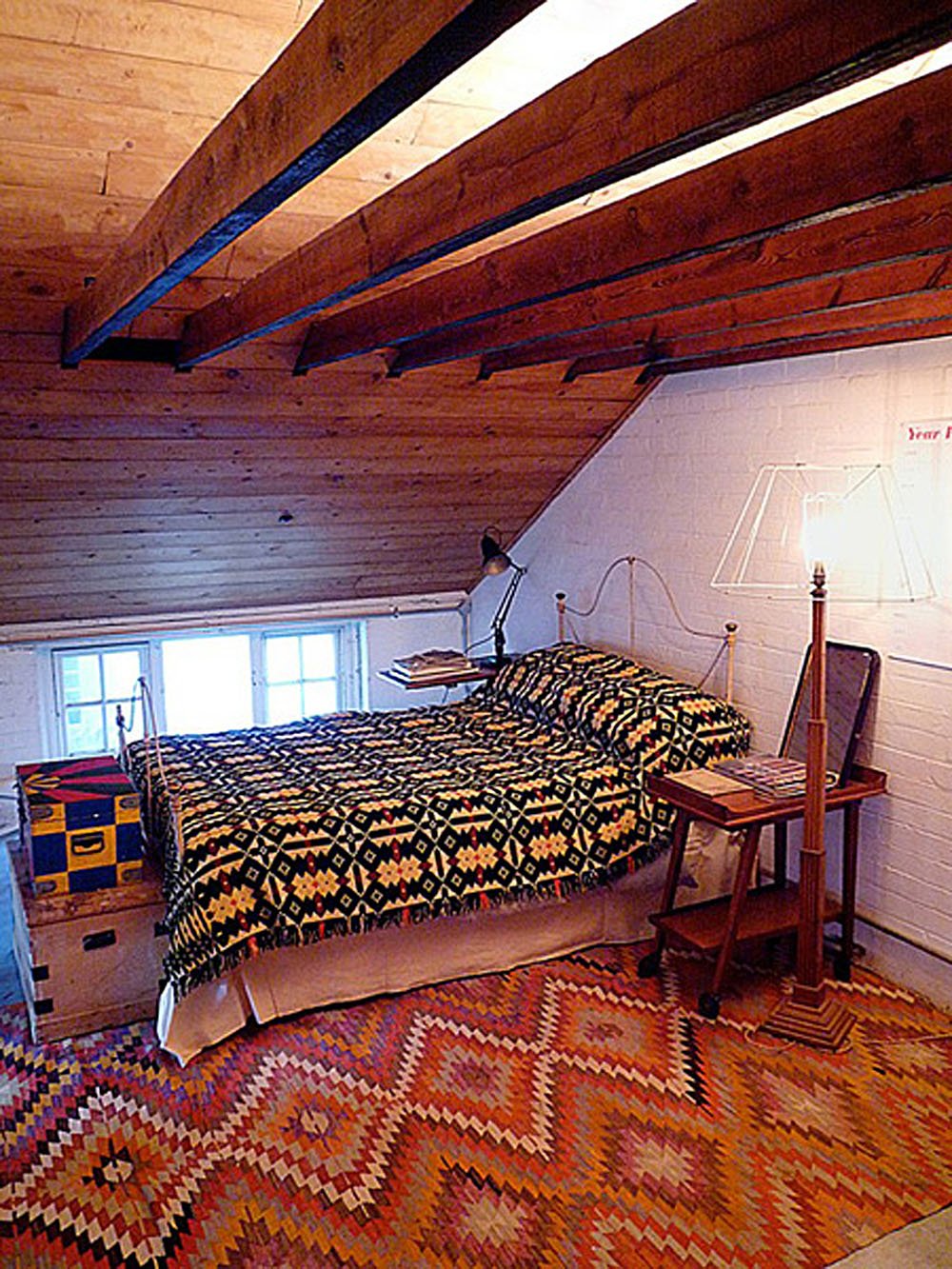 A cozy attic bedroom with a sloped wooden ceiling, a bed with a black and yellow patterned quilt, a small window, a wooden side table, and a tall wooden standing lamp.