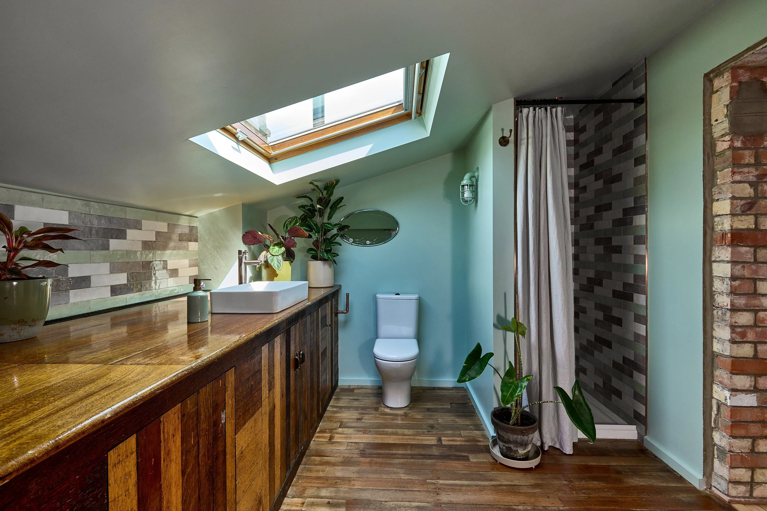 Bathroom with skylight, wooden cabinets, a tile wall, a white sink, plants, a toilet, and a shower area with curtain and brick wall.