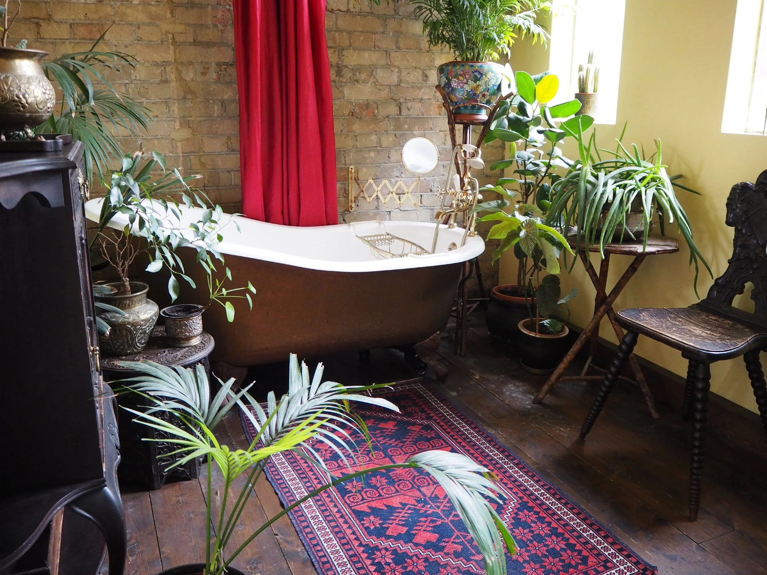 Vintage bathroom with clawfoot tub, multiple potted plants, antique furniture, a red curtain, and a patterned rug on wooden floors.