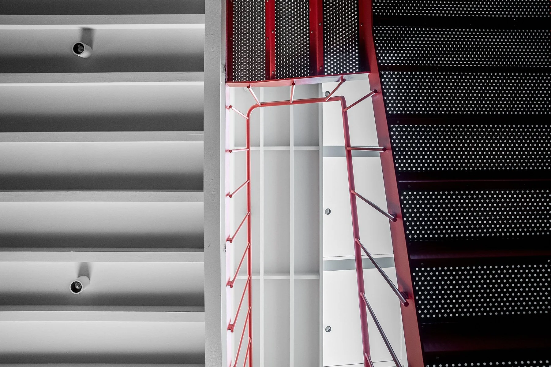 A photograph taken from below showing a multi-level indoor staircase with black, red, and white railings and steps, and security cameras on the ceiling.