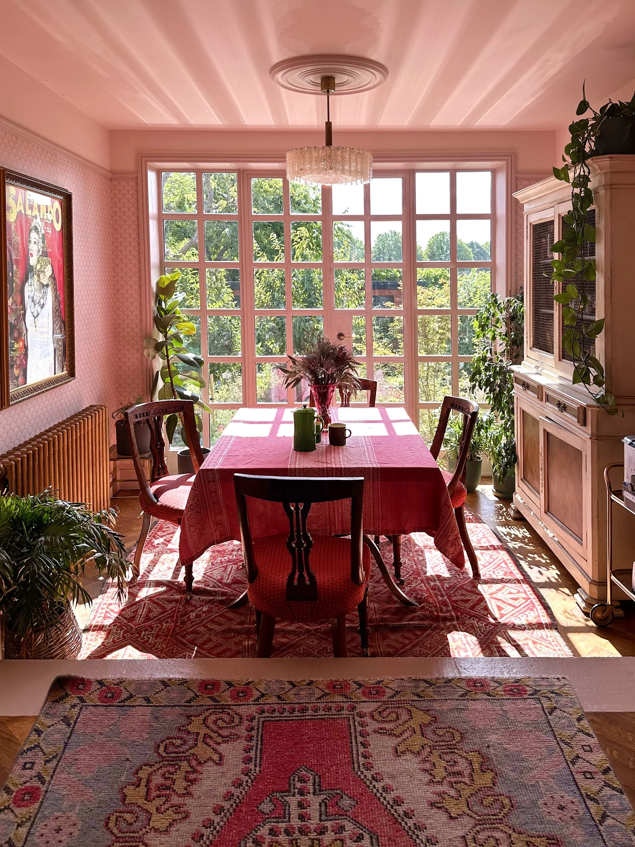 Bright dining room with large window, pink walls, a red tablecloth, and surrounded by wooden chairs. Decor includes plants, a vintage picture on the wall, and a wooden cabinet.
