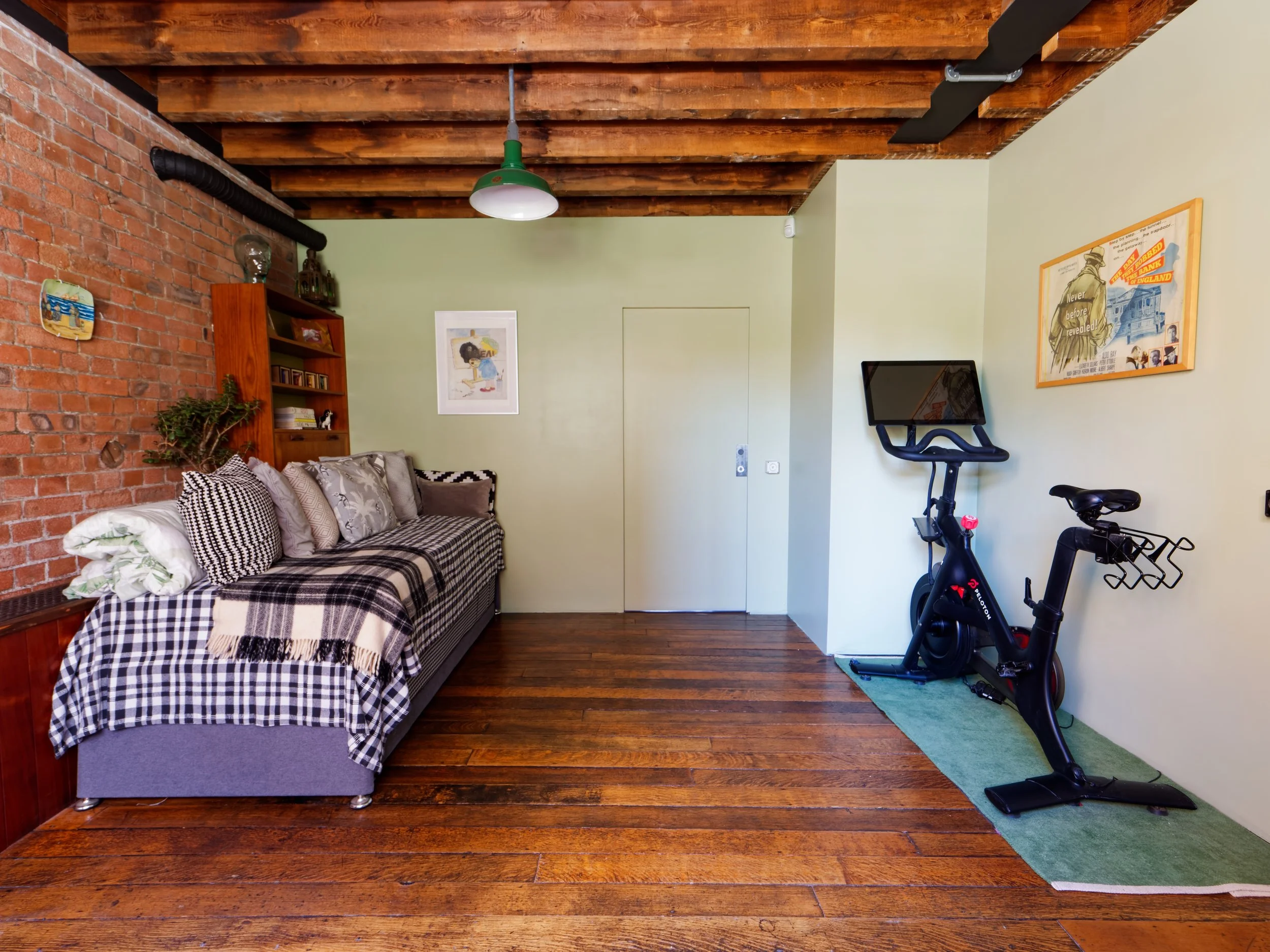 Living room with green walls, exposed brick wall, wooden ceiling, hardwood floor, a gray sofa with checkered blanket and multiple pillows, a tall wooden bookshelf with decor and books, a framed artwork on the wall, a stationary exercise bike with a m