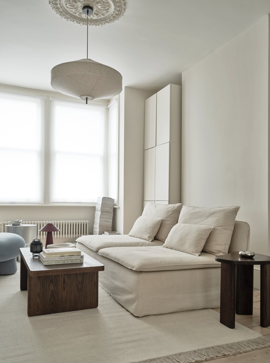 Bright living room with a beige sofa, dark wooden side tables, a stack of books, and large windows with white blinds, creating a minimalistic and airy atmosphere.