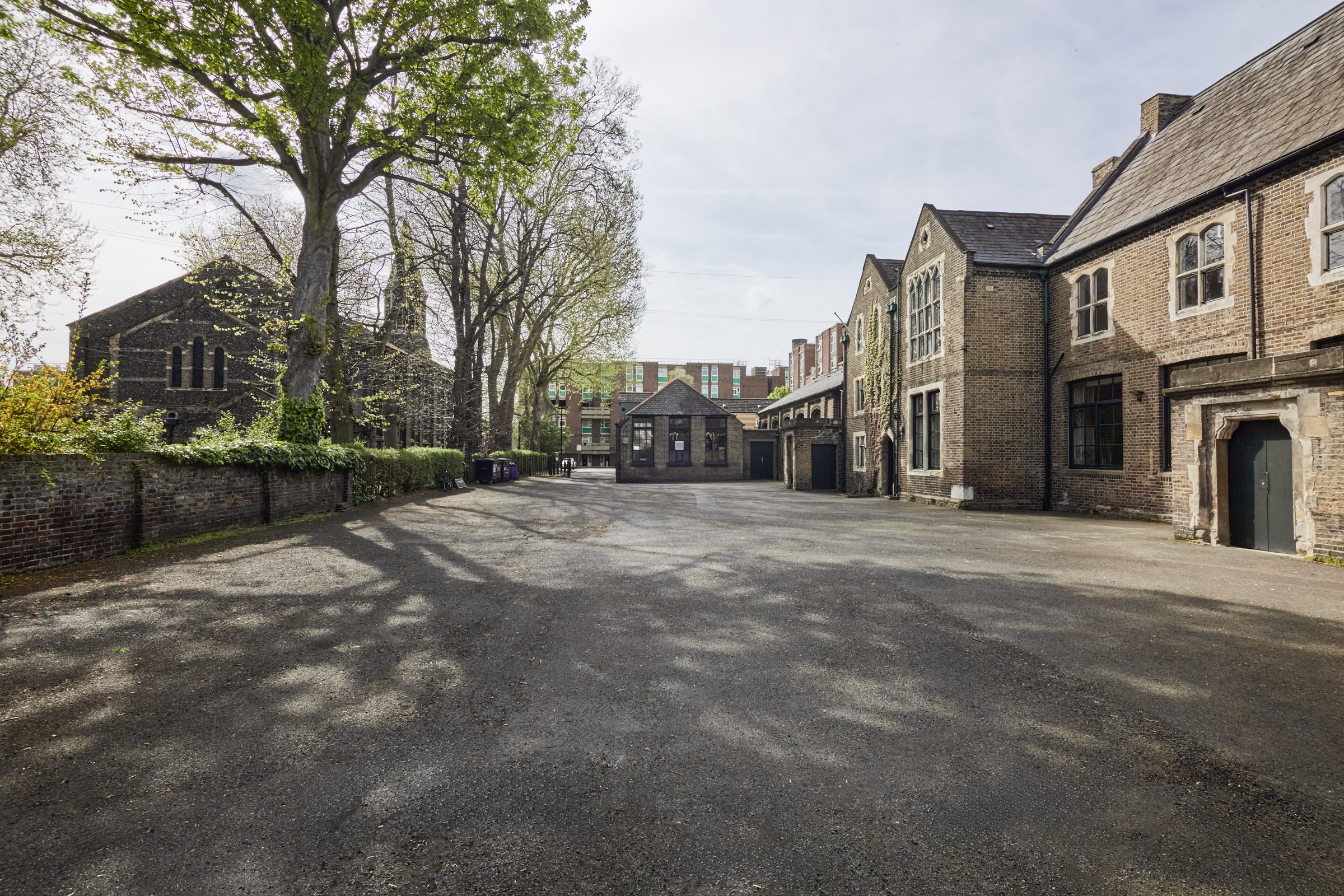 An empty gravel yard surrounded by brick buildings with trees on the left and more distant buildings in the background.