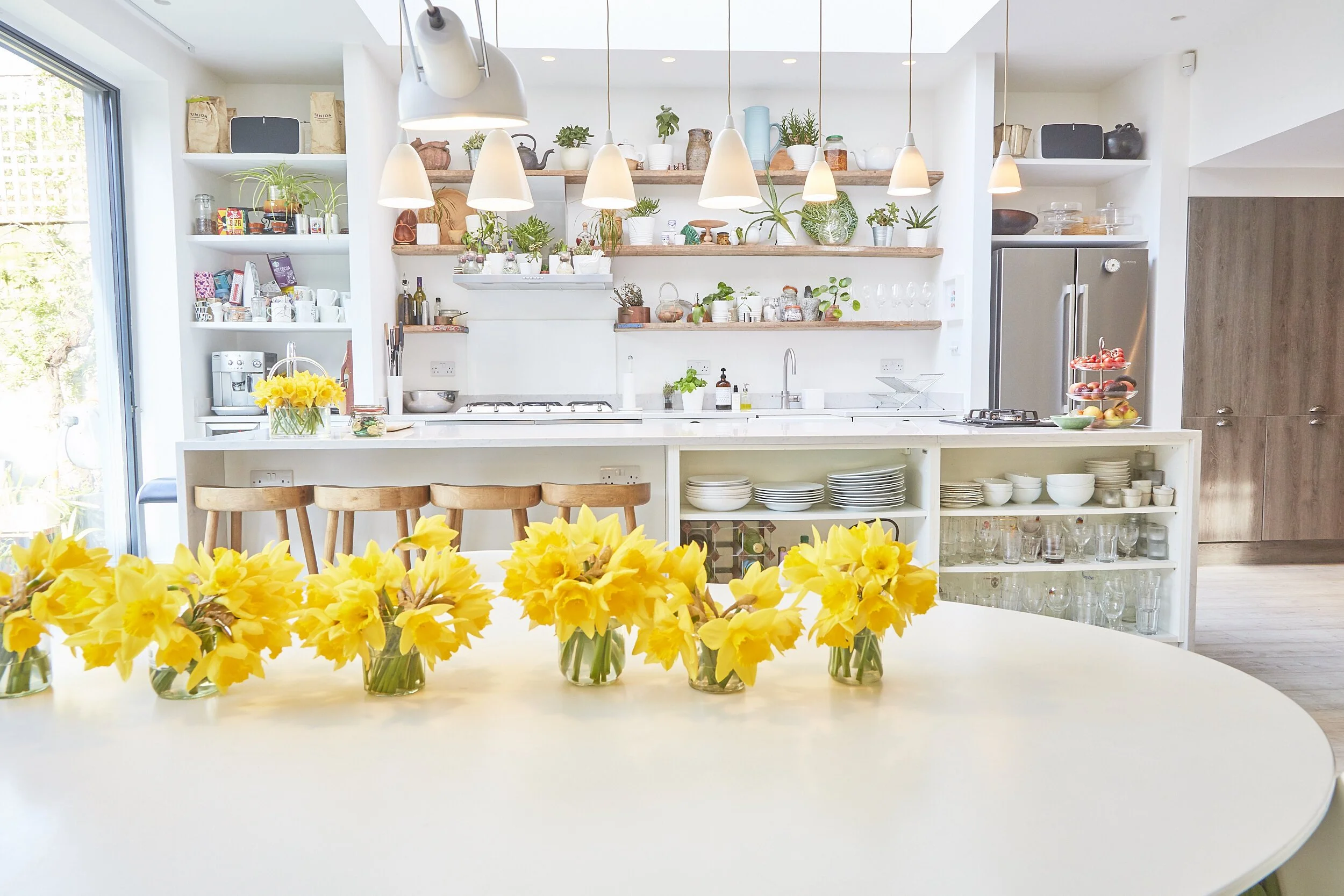 Bright, modern kitchen with white counters, open shelving with plants and kitchenware, a large refrigerator, and a round table with multiple glass vases filled with yellow flowers.