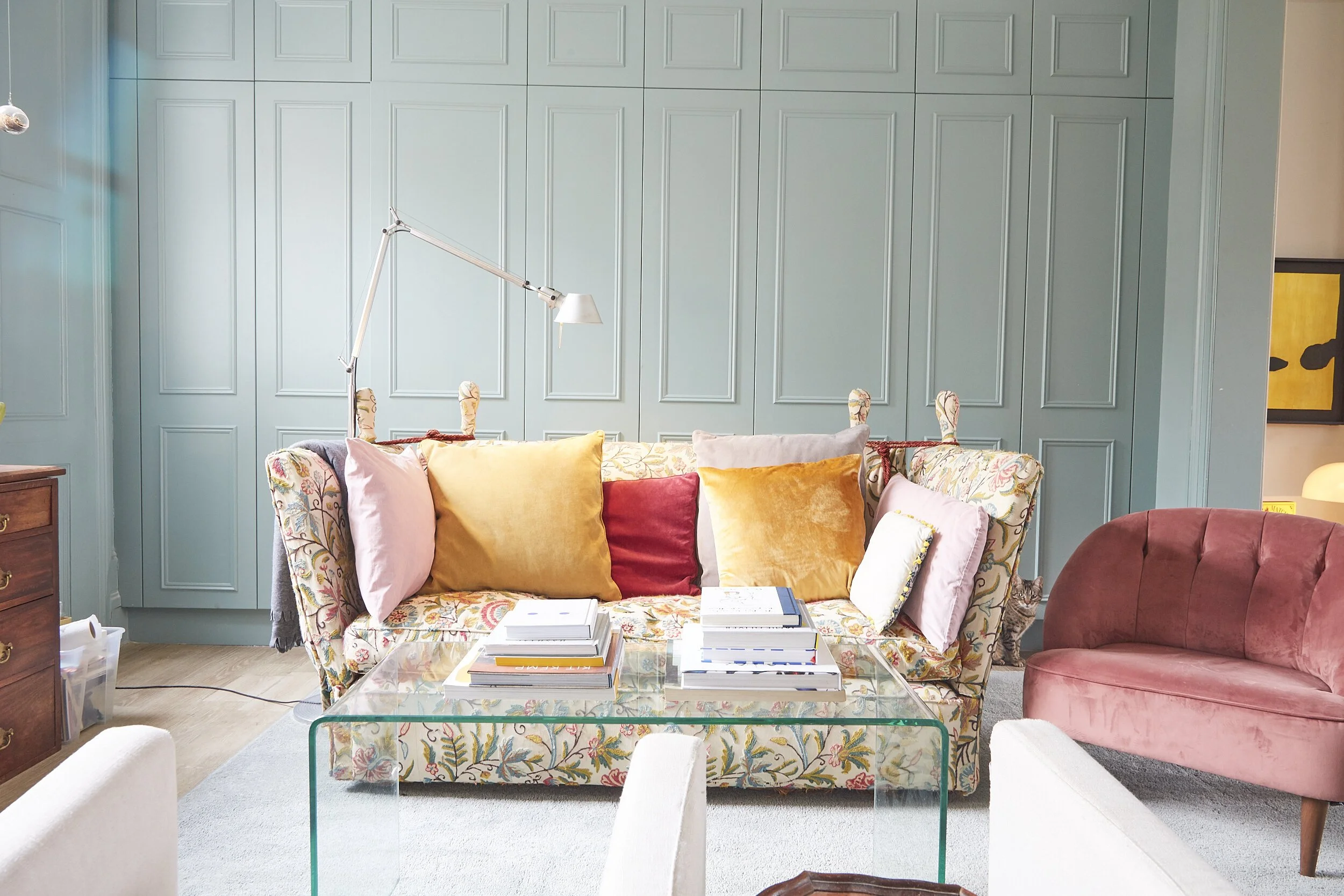A living room with a blue paneled wall, floral-patterned sofa with various colored pillows, a glass coffee table with stacks of books, a pink velvet armchair, and a small tabby cat peeking from behind the sofa.