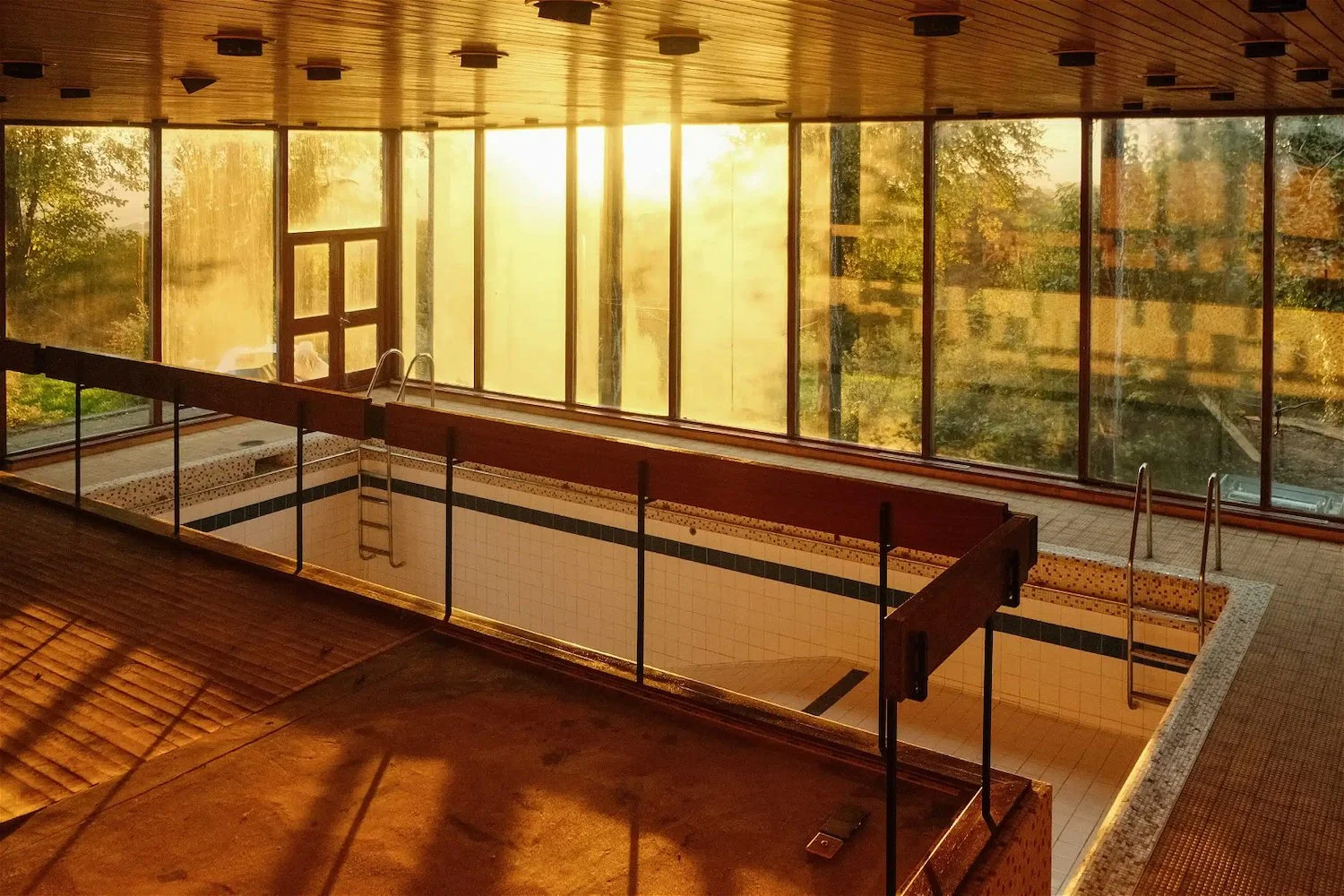 Indoor swimming pool area with large glass windows showing trees outside, sunlight streaming in, and a wooden ceiling.