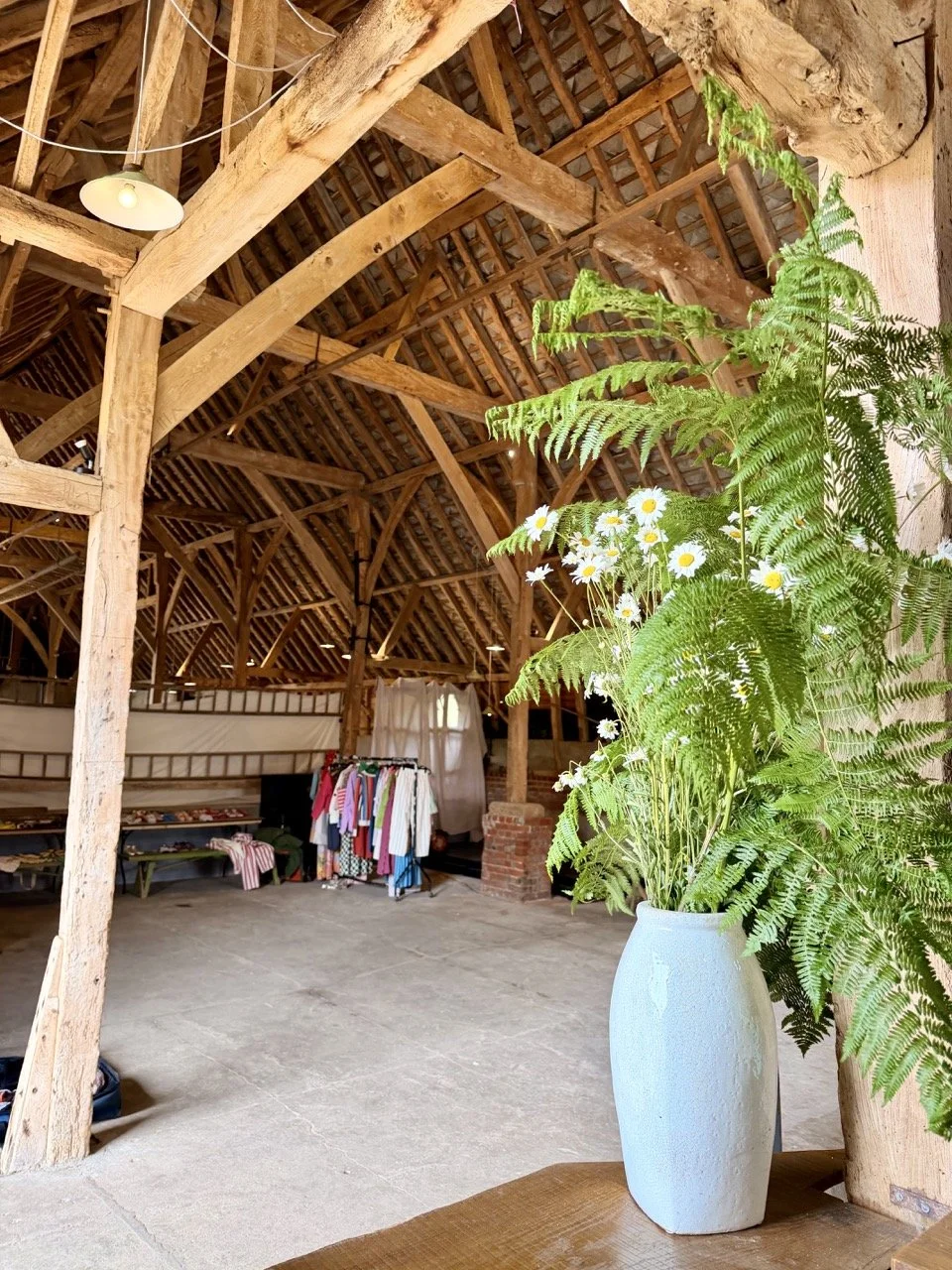 A white vase with green fern leaves and white daisies sits on a wooden surface inside a rustic barn with exposed wooden beams and a tiled roof.