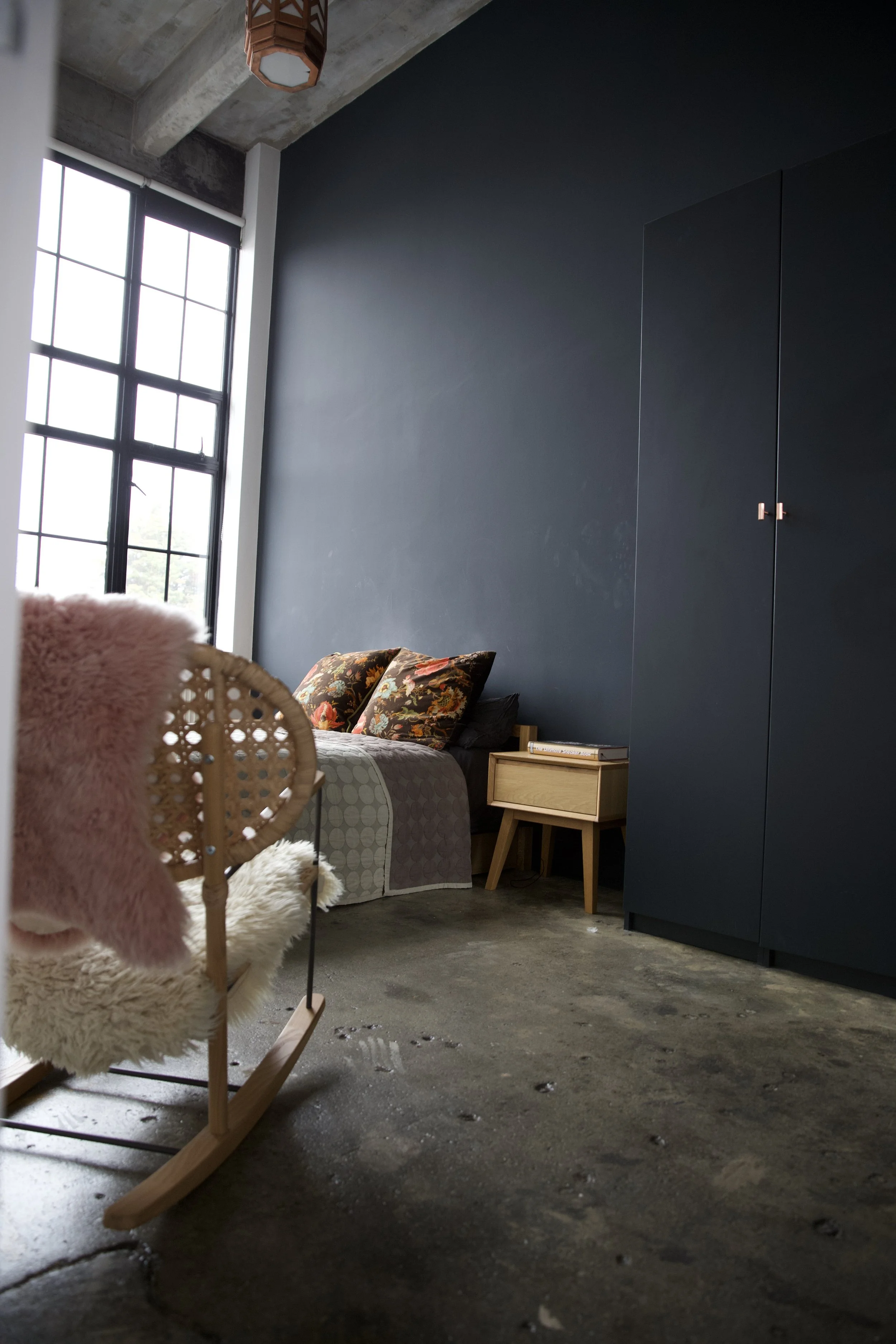 Interior of a modern bedroom with dark blue walls, large window, bed with floral pillows, wooden nightstand, wardrobe, pink and white plush rocking chair, and concrete floor.