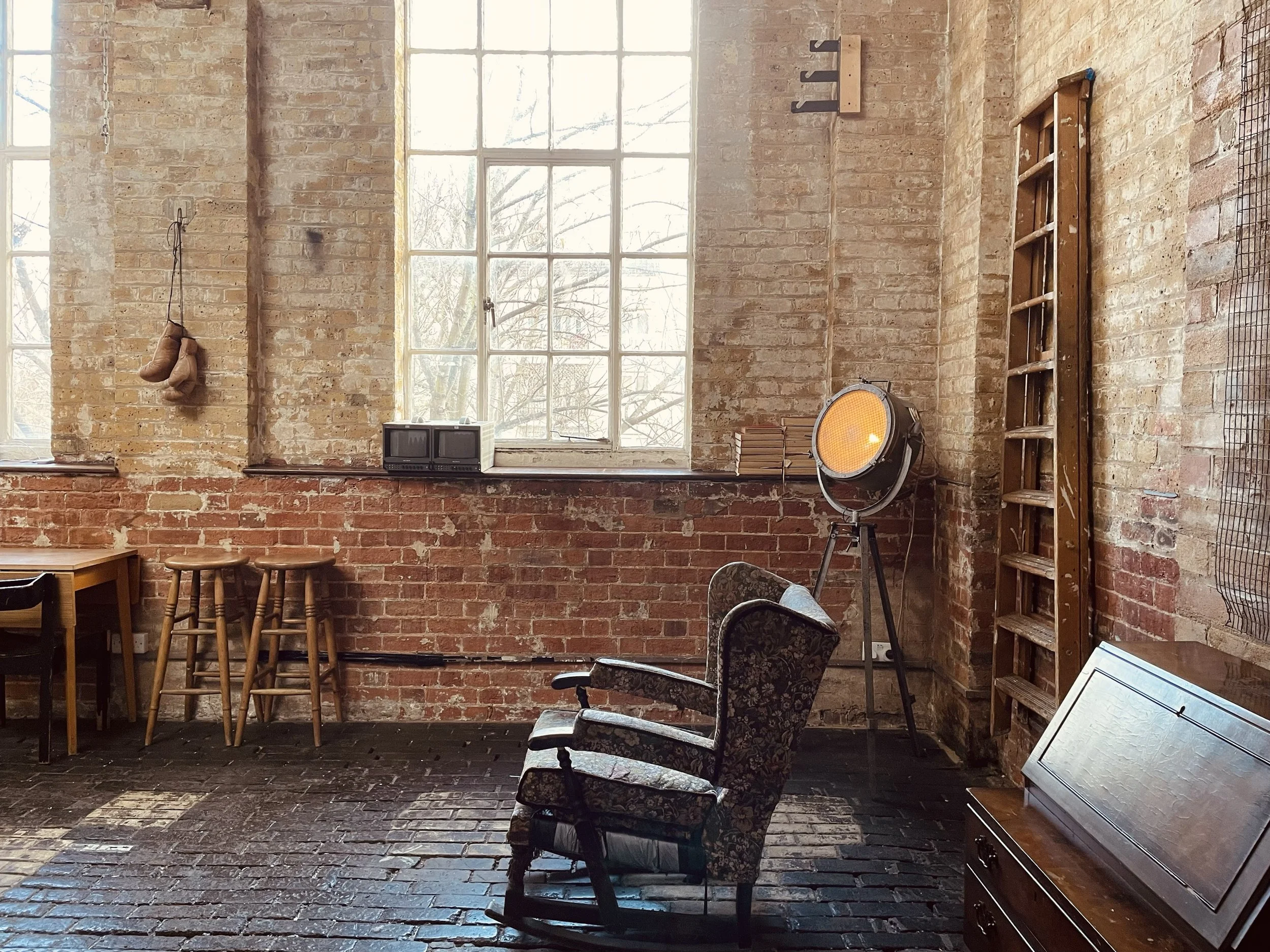 Room with brick walls and large window. Includes vintage armchair, wooden table and stools, ladder, ladder, spotlight, and small TV.
