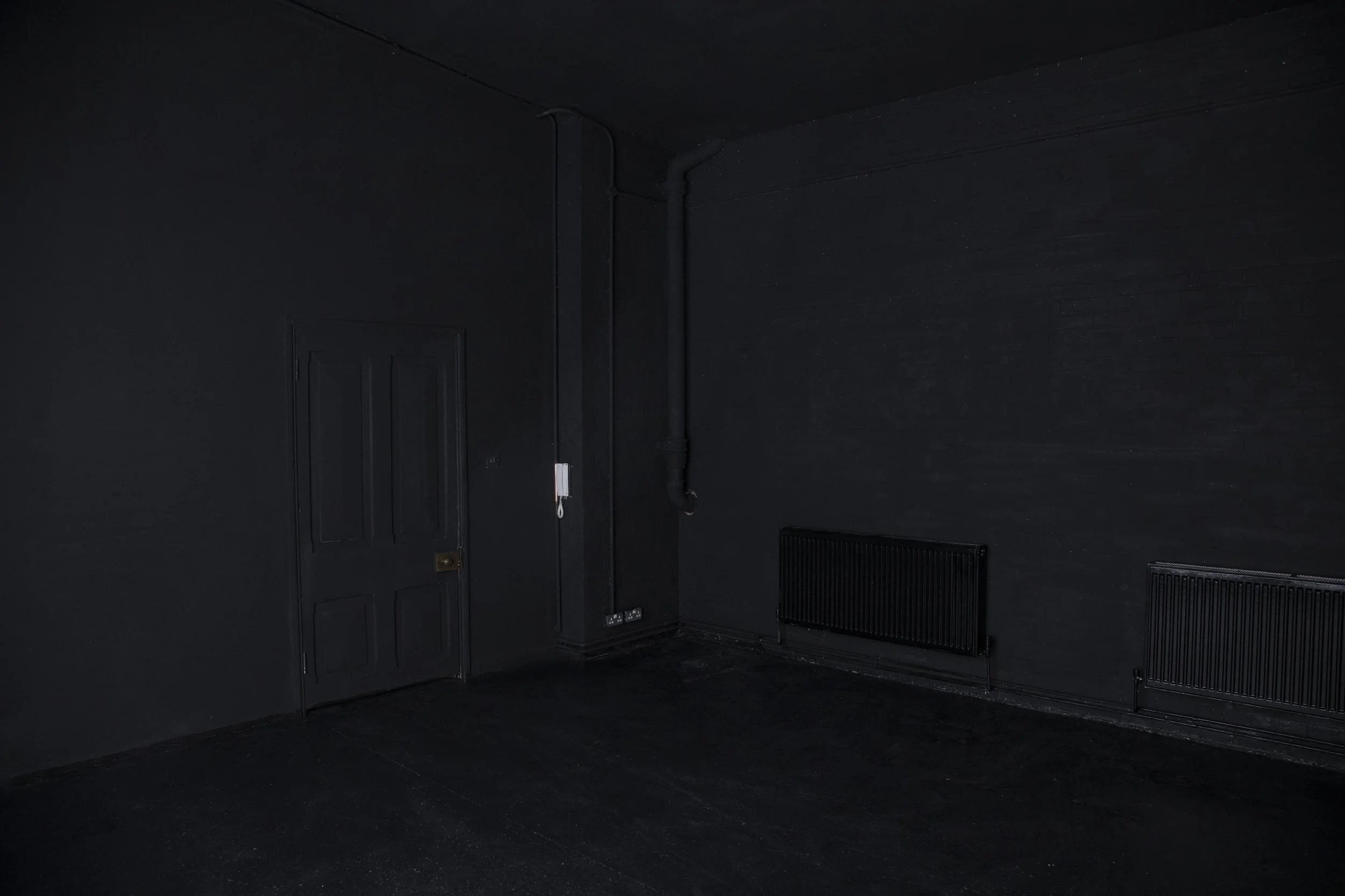 Dark, empty room with black walls, black floors, a black door, a white intercom on the wall, exposed pipes, and black radiators.