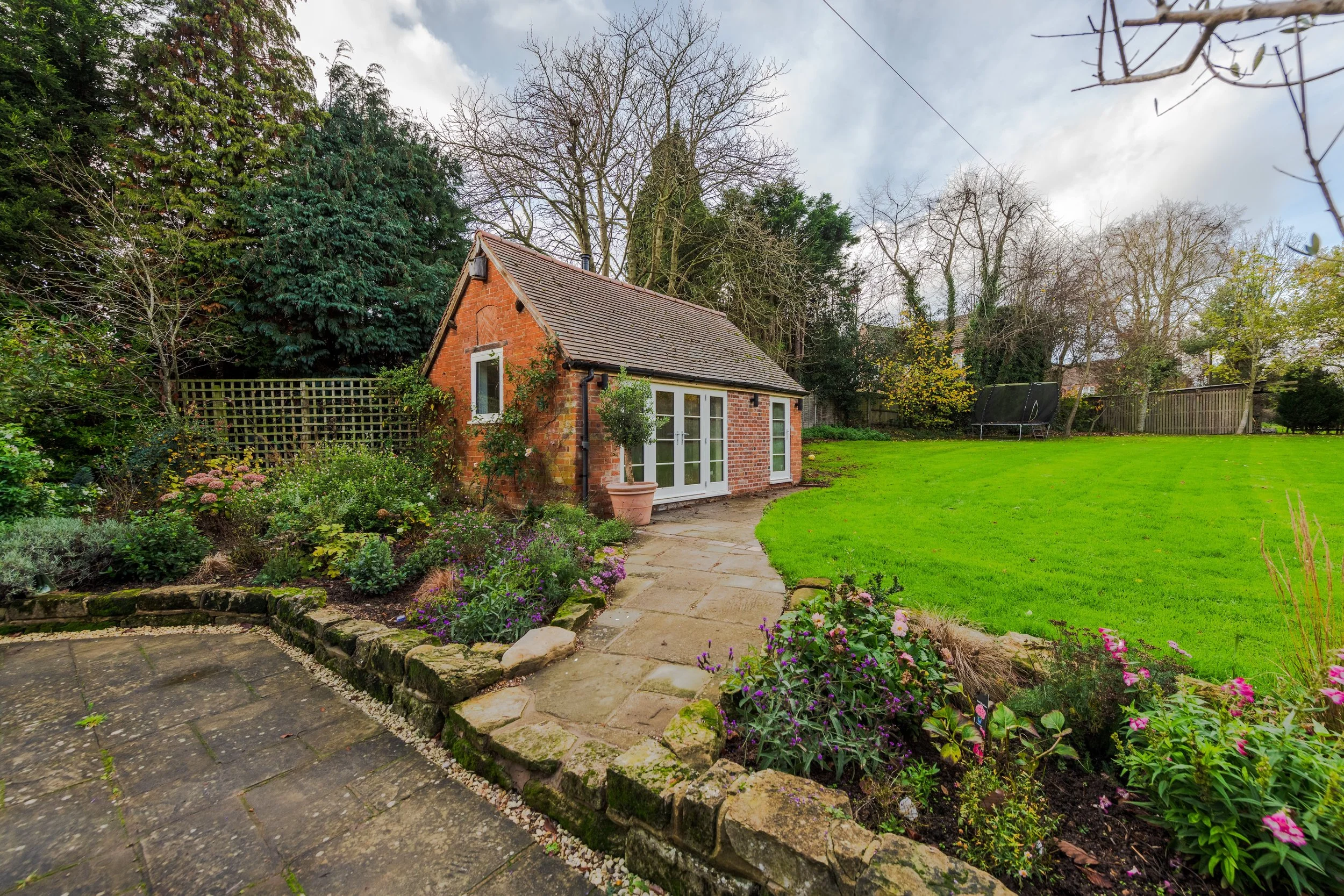A small brick garden house with French doors, a pitched roof, and a potted topiary outside, set in a lush, landscaped backyard with a well-maintained lawn, colorful flowers, trees, and a trampoline in the background.