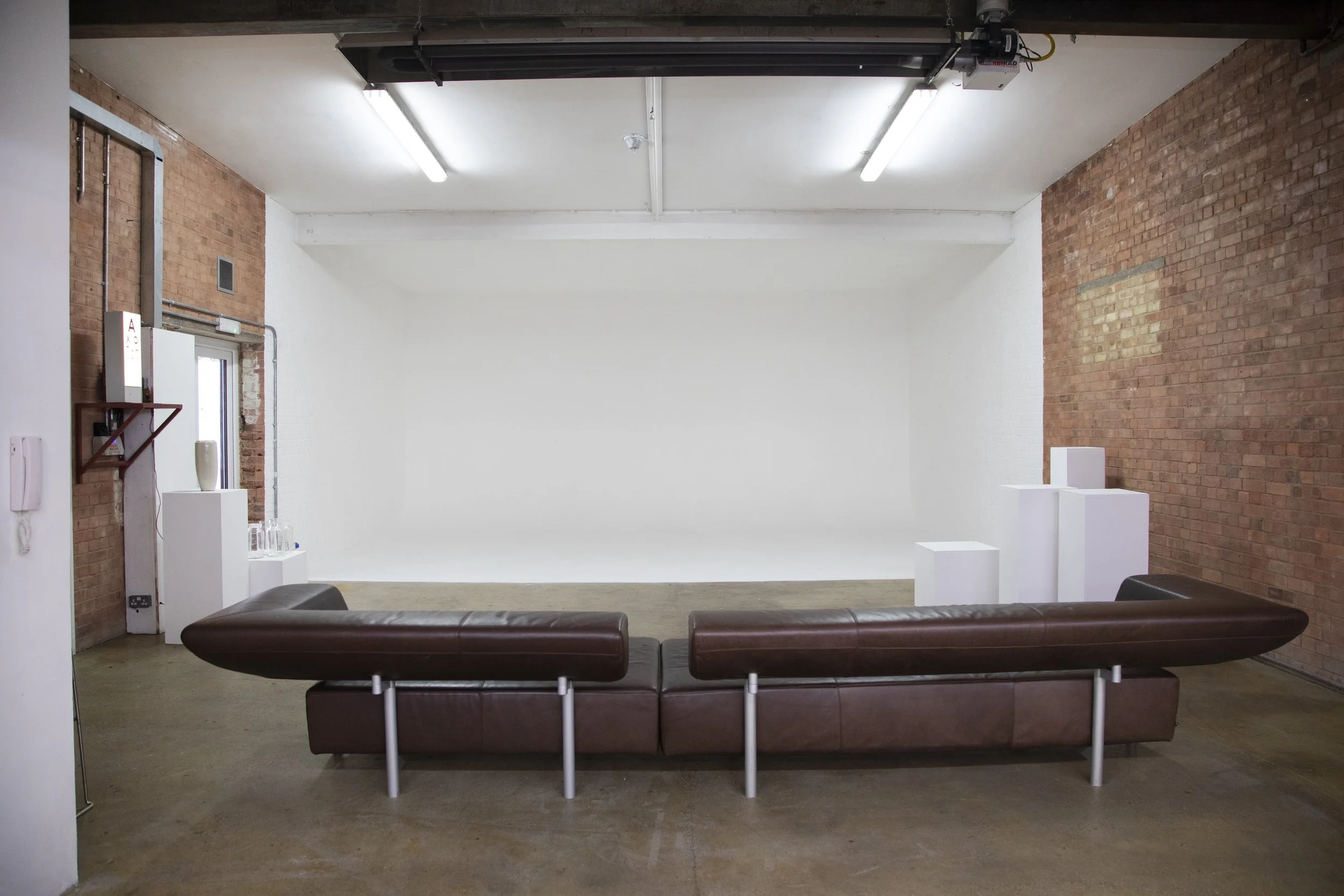 Empty gallery or studio space with white walls, exposed brick walls, ceiling-mounted lights, a brown leather sofa, and white display pedestals, some with glassware.