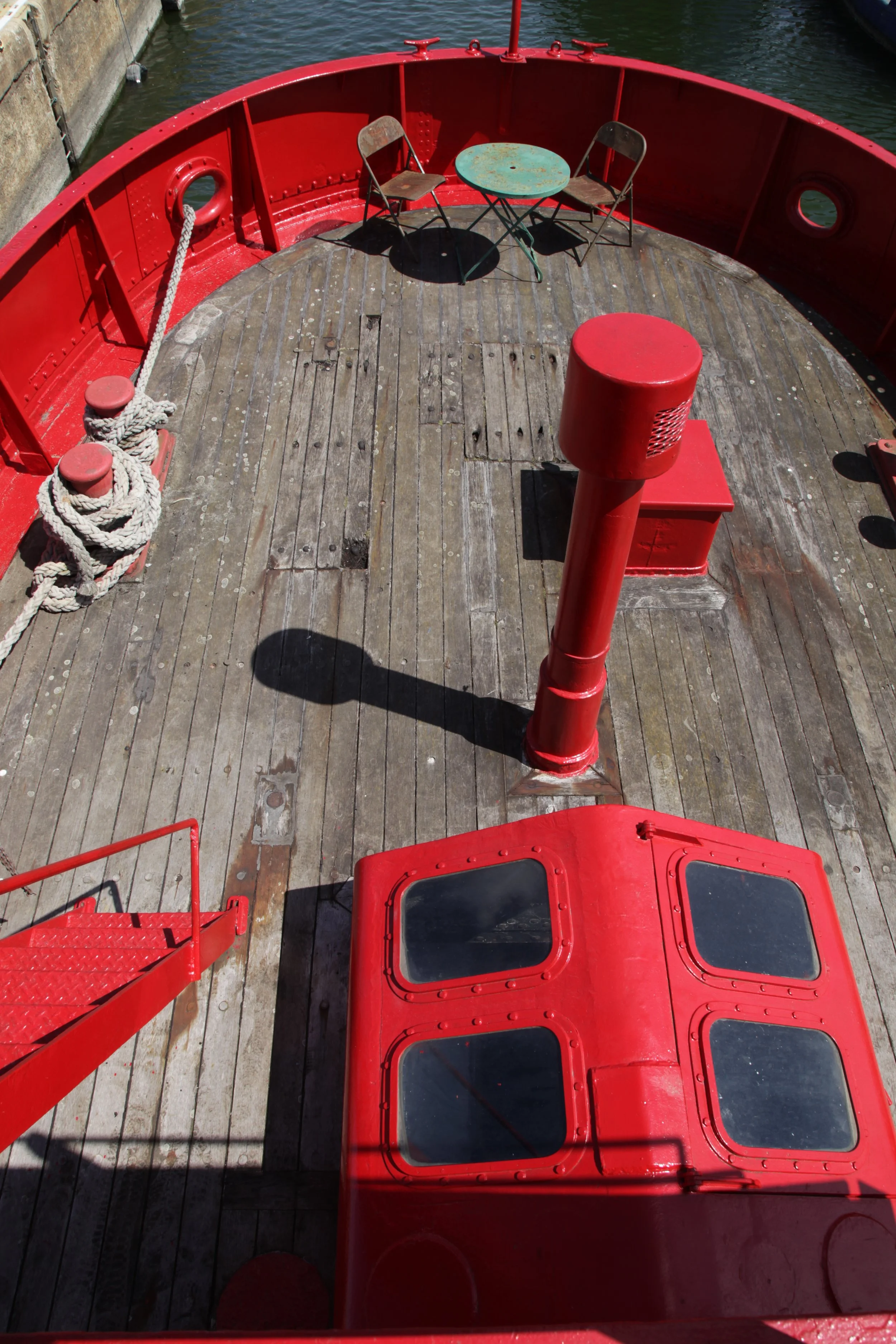 View of the deck of a red boat with a small green table and two chairs, along with coiled ropes and a red hatch with windows.