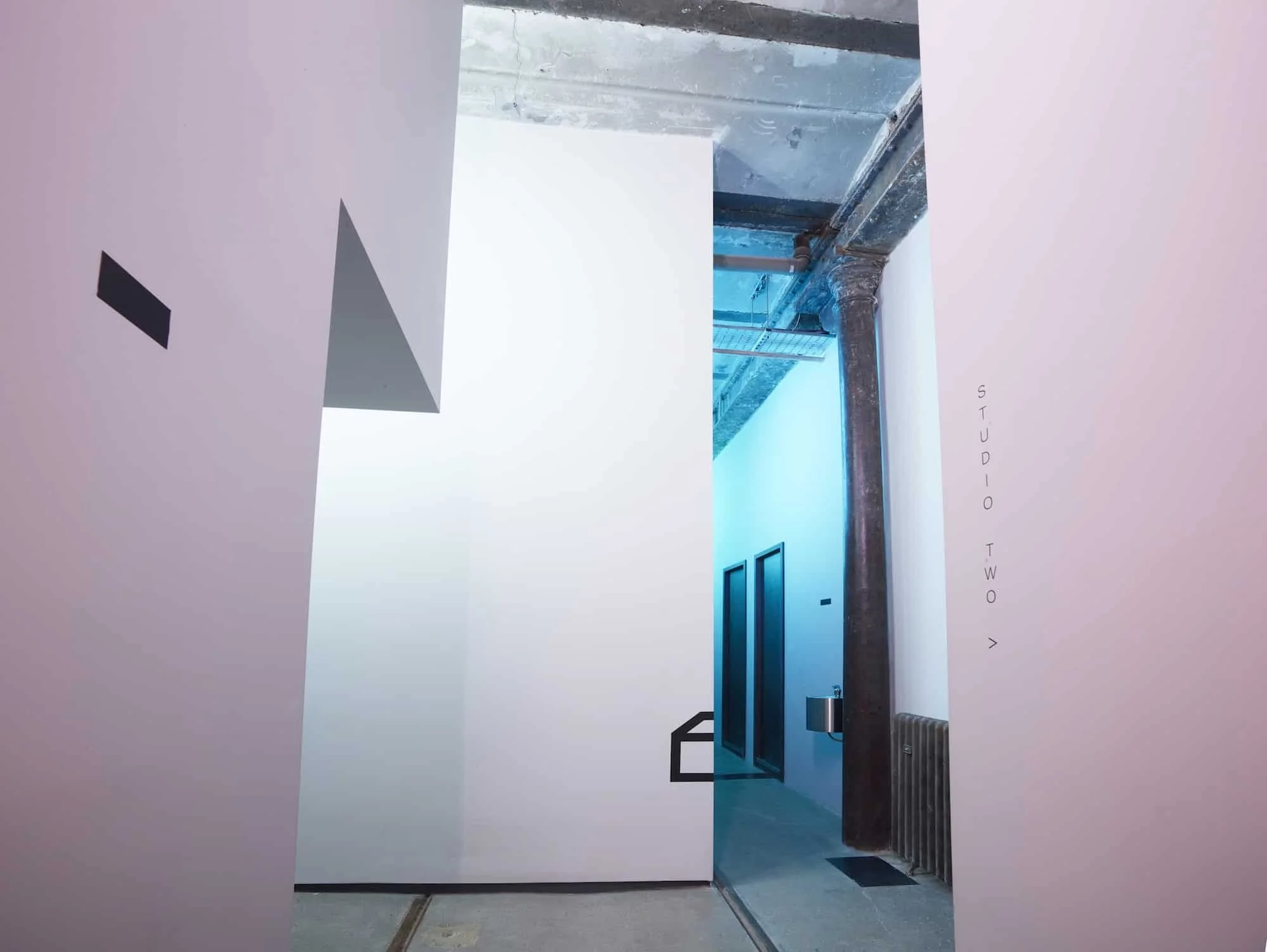 Interior of an art gallery or museum with white walls, featuring a clear blue wall with two black-framed windows or artworks, a dark column, and a visible exhaust pipe. The right wall has vertical text that says 'STUDIO TWO' with an arrow pointing ri