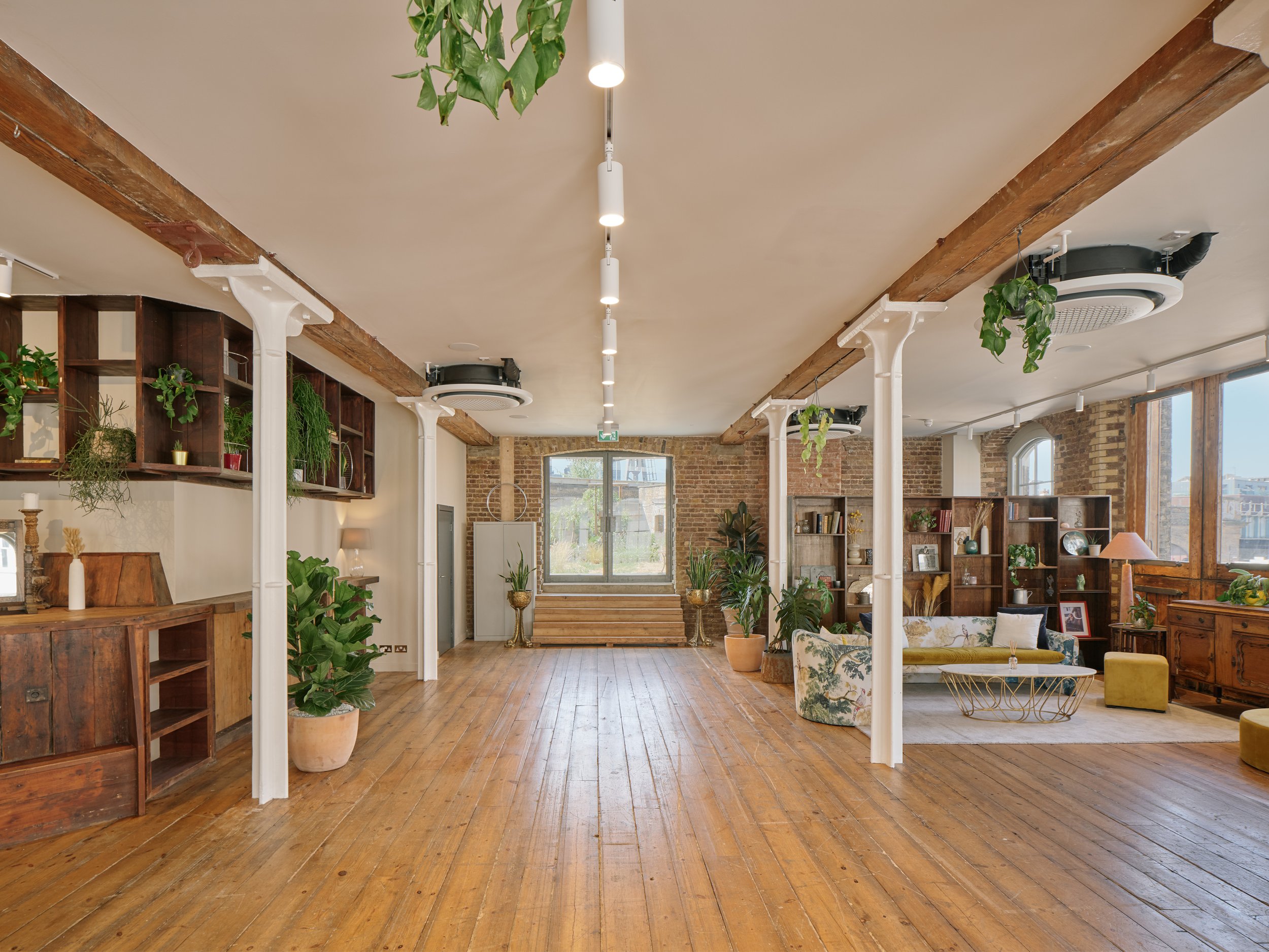 Spacious living room with wooden floors, brick walls, large windows, green plants, and vintage furniture.